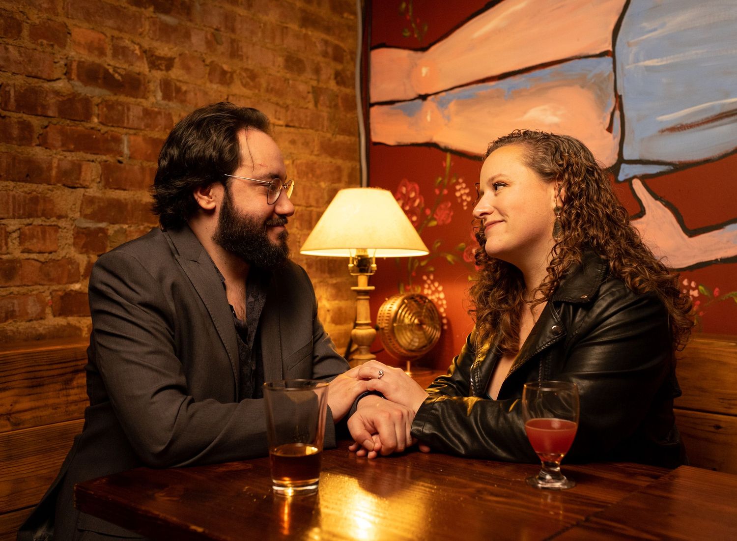 Man and woman holding hands at a bar table with warm lamp light between them.