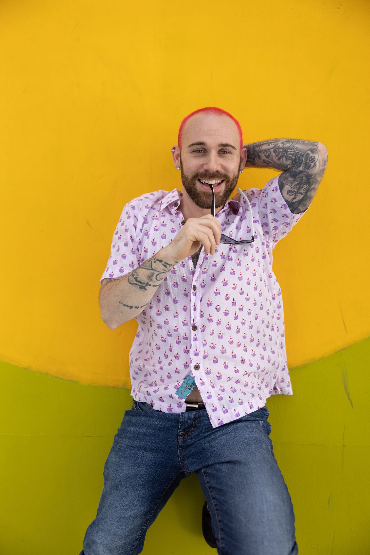 Smiling tattooed man with pink hair holding glasses against vibrant yellow wall wearing patterned shirt and jeans.