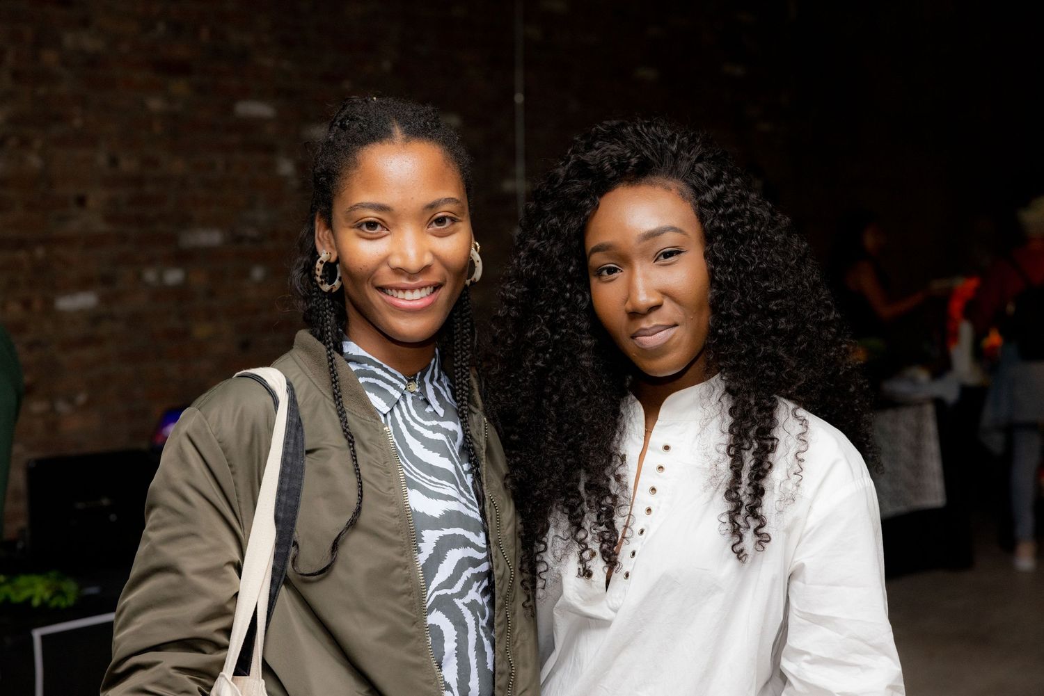 Two young Black women smiling together at an indoor social gathering event.