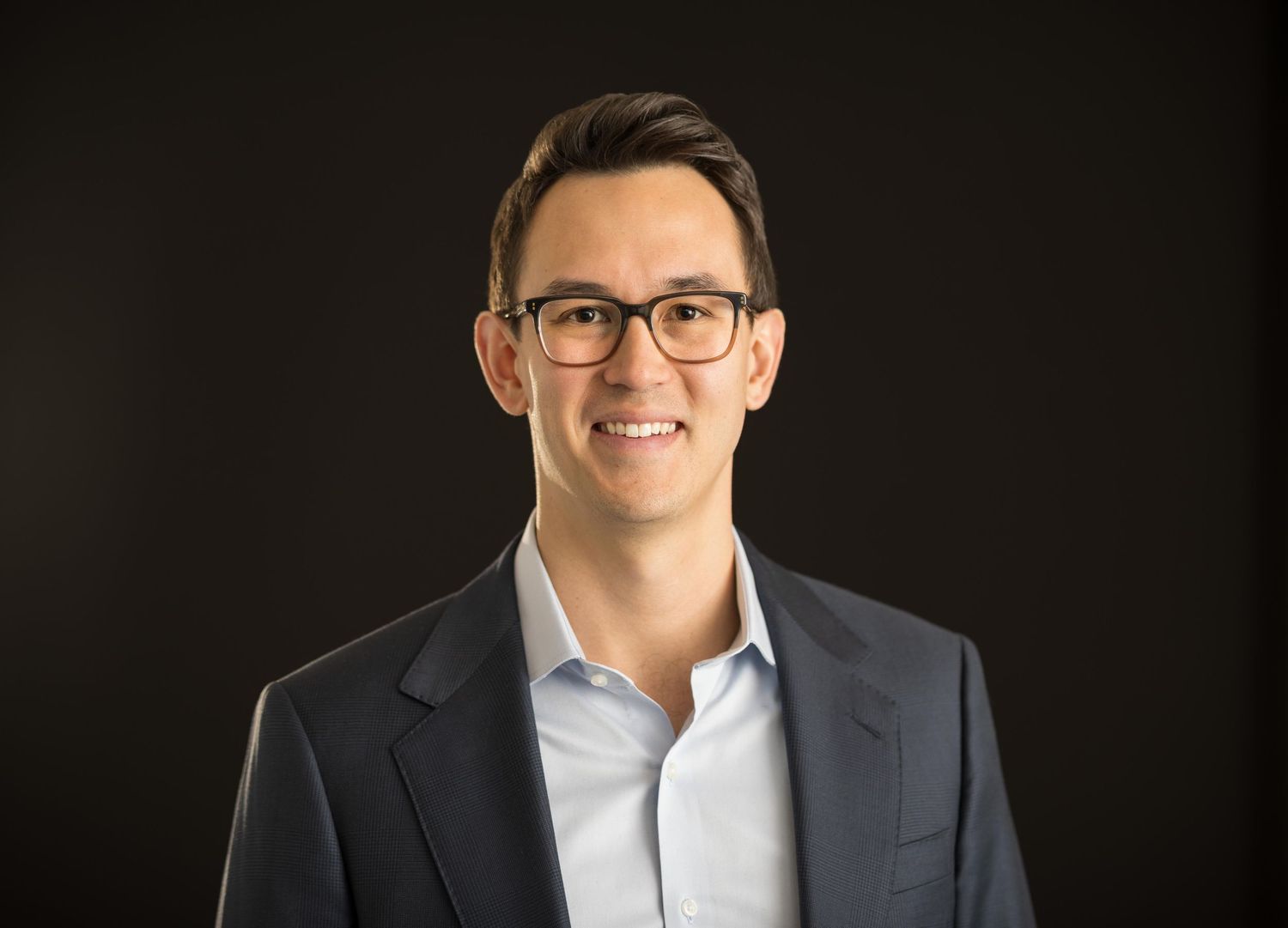 Professional headshot of a smiling man wearing glasses and a navy blazer against a dark background.