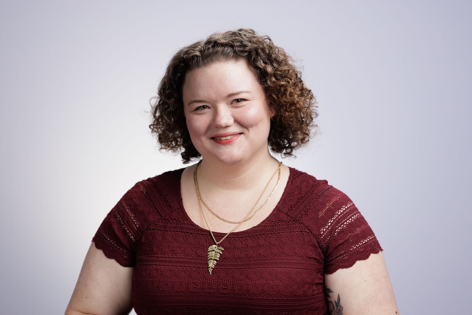 Woman with curly hair smiling, wearing a red lace top and gold necklace, professional headshot on gray background.