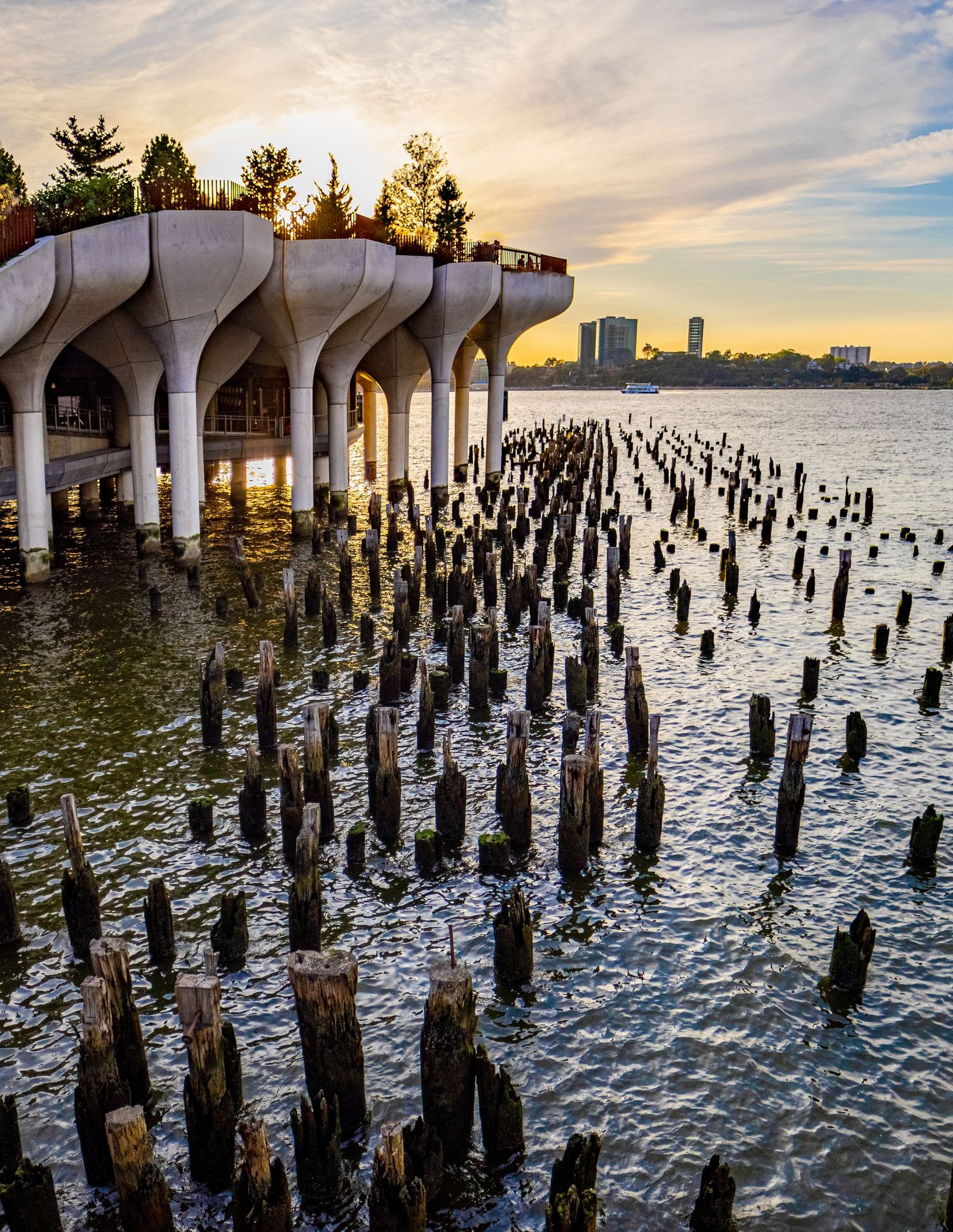 Sunset view of Little Island park on the Hudson River with weathered wooden pier pilings in the foreground.