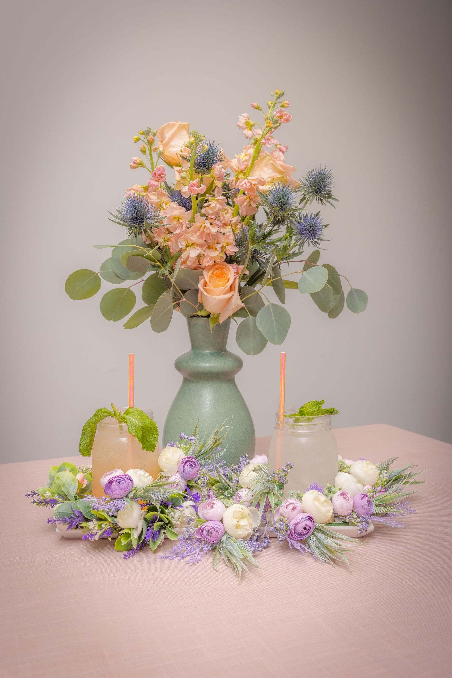 Elegant spring wedding table centerpiece with peach roses, blue thistles, and purple ranunculus in mint vase.