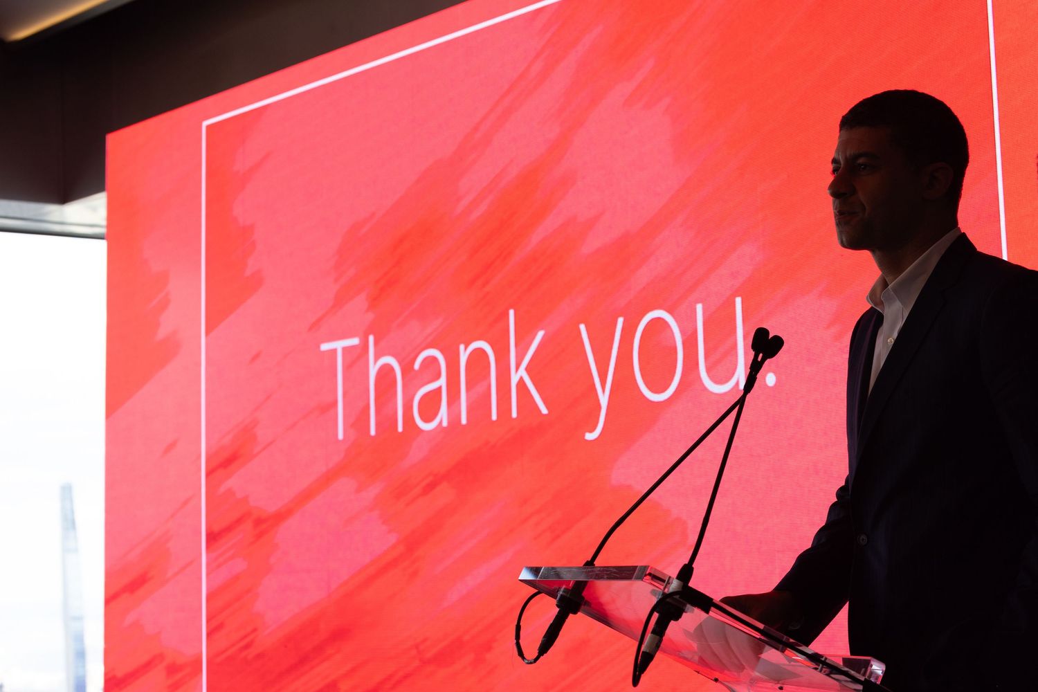 Male speaker silhouetted against a red Thank You slide at a corporate presentation podium.