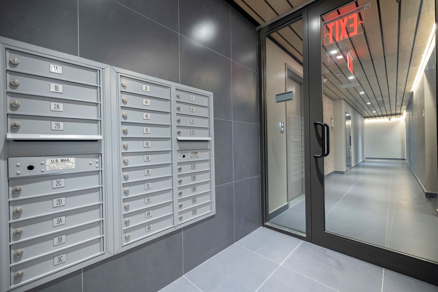 Modern building mailroom with rows of metal mailboxes, gray tile walls, and a glass corridor door.