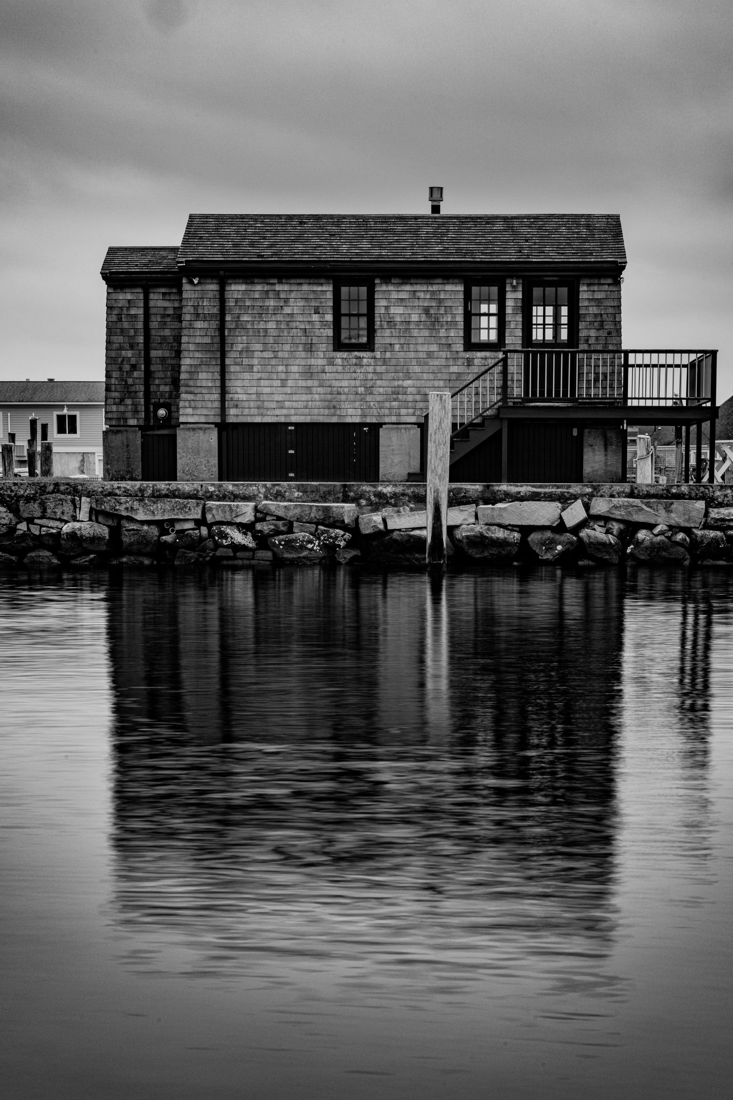 Black and white photo of a waterfront shingled house reflecting in calm harbor waters under overcast sky.
