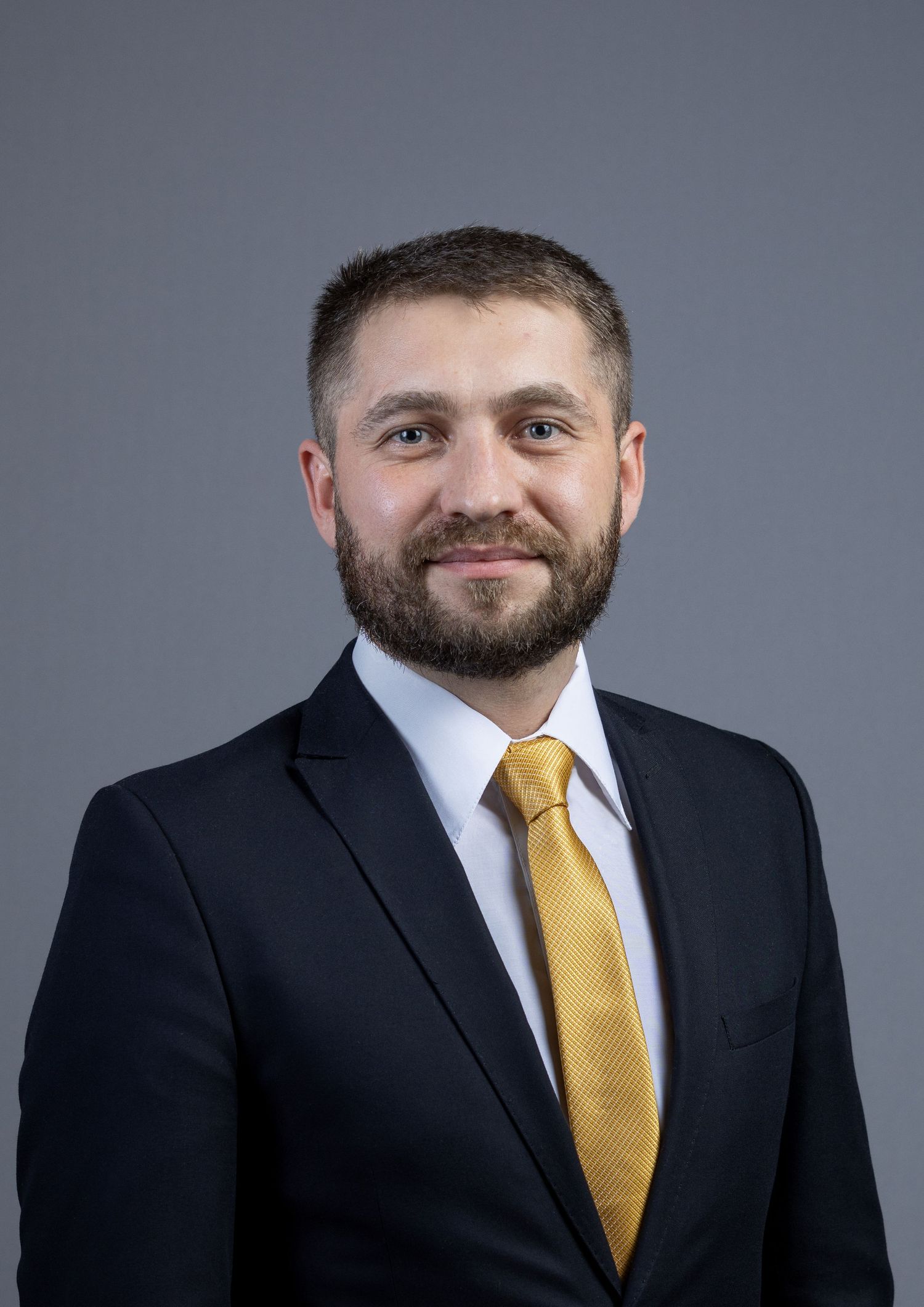 Professional man with beard wearing black suit and gold tie against gray background.
