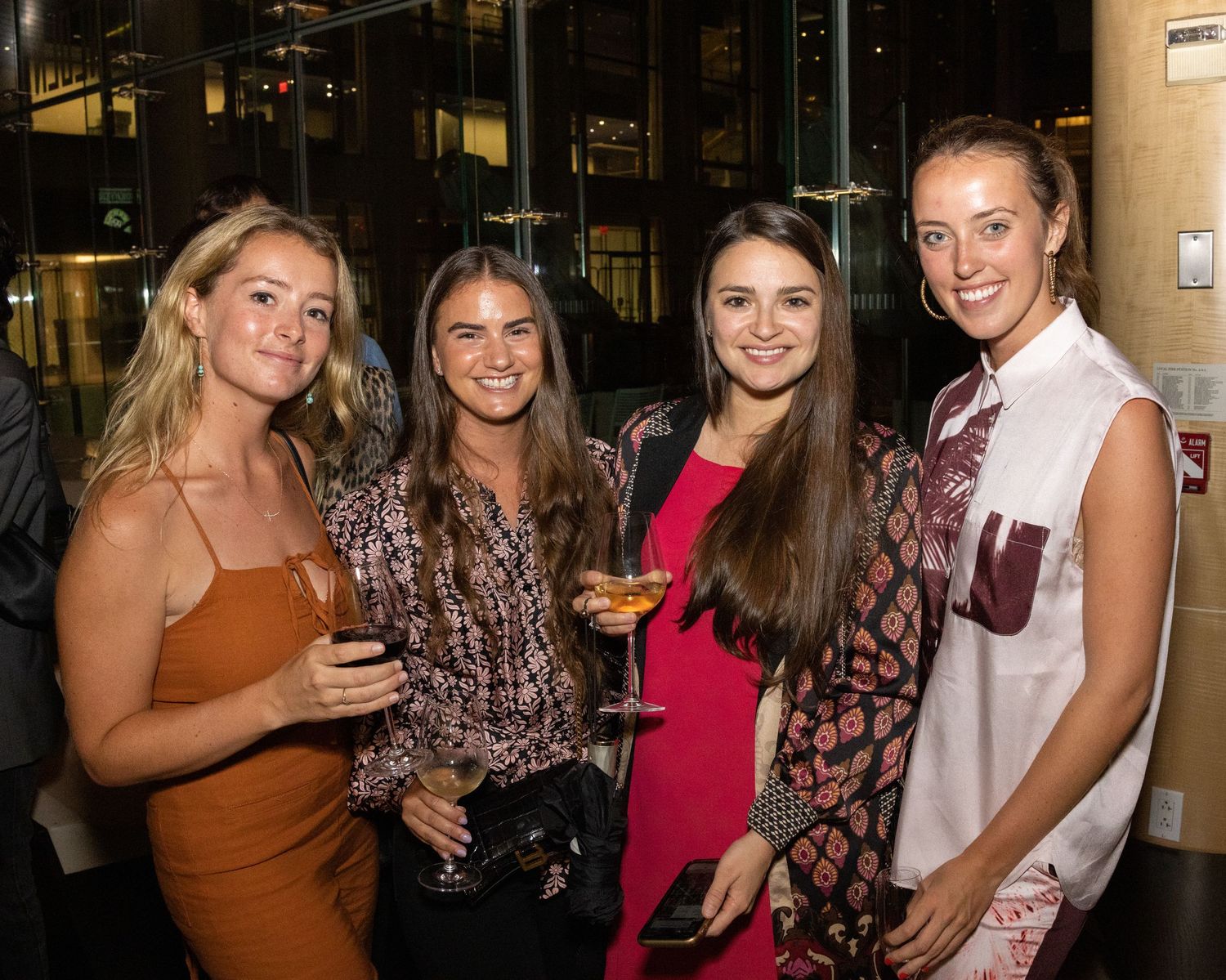 Four smiling white women holding cocktails posing together at an indoor evening social gathering event.