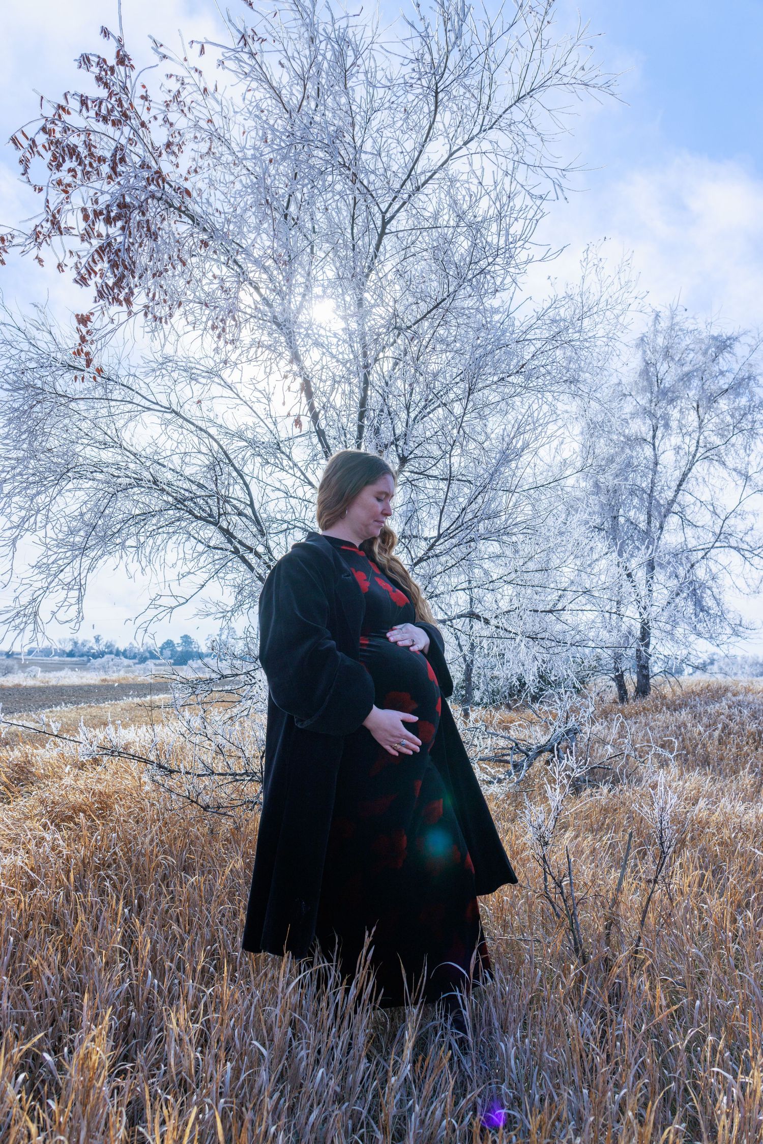 Pregnant woman in a black coat and red dress cradling her belly in a snowy winter landscape.