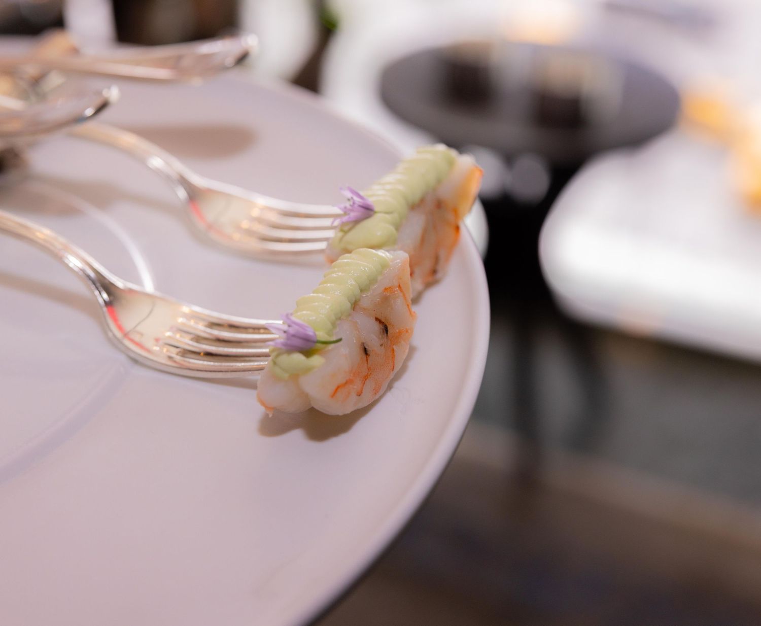 Elegant shrimp appetizers served on silver forks atop a white plate at a fine dining event.