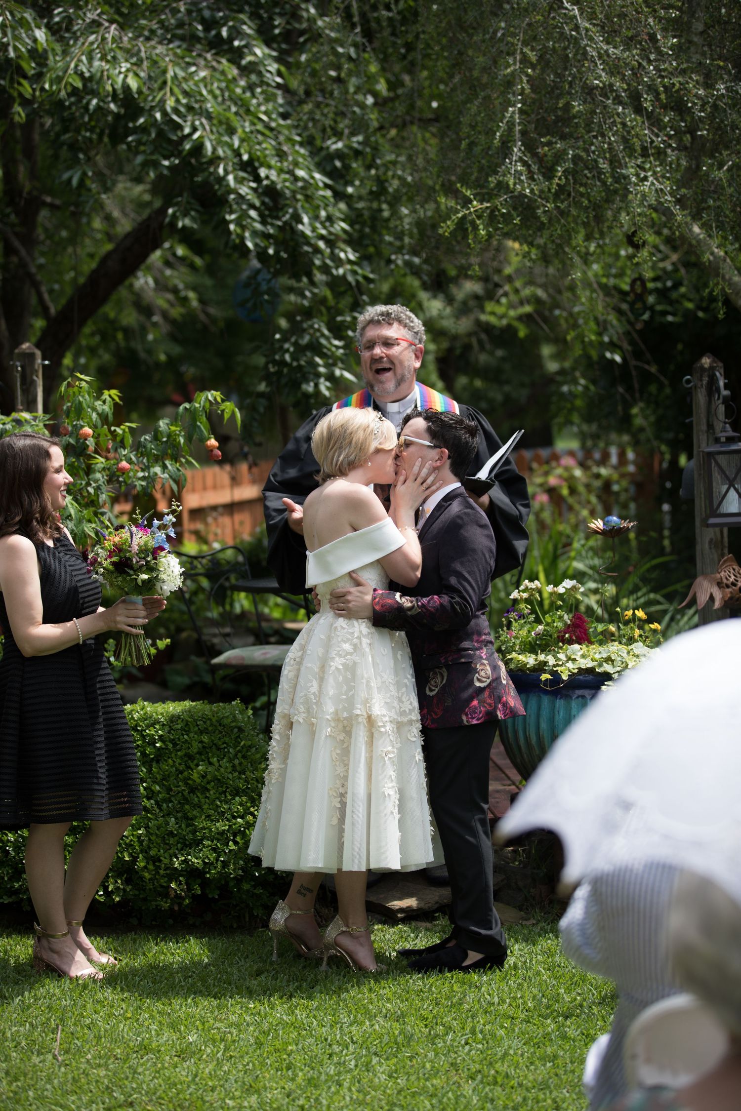 Two brides share their first kiss at an outdoor garden wedding ceremony with officiant and guests.