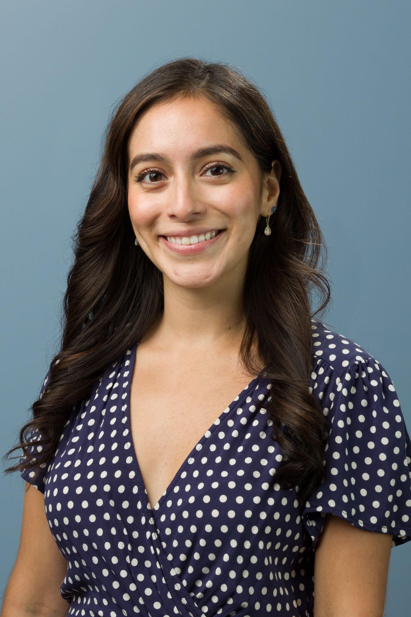 Smiling young woman with dark wavy hair wearing a navy polka dot dress in a professional headshot.