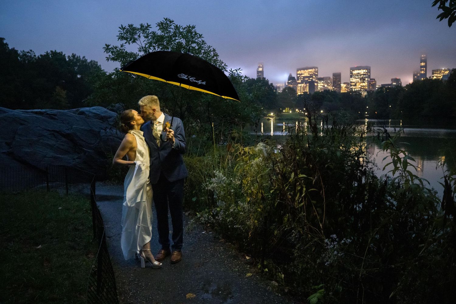 Wedding couple kissing under black umbrella near Central Park lake with illuminated NYC skyline at night.