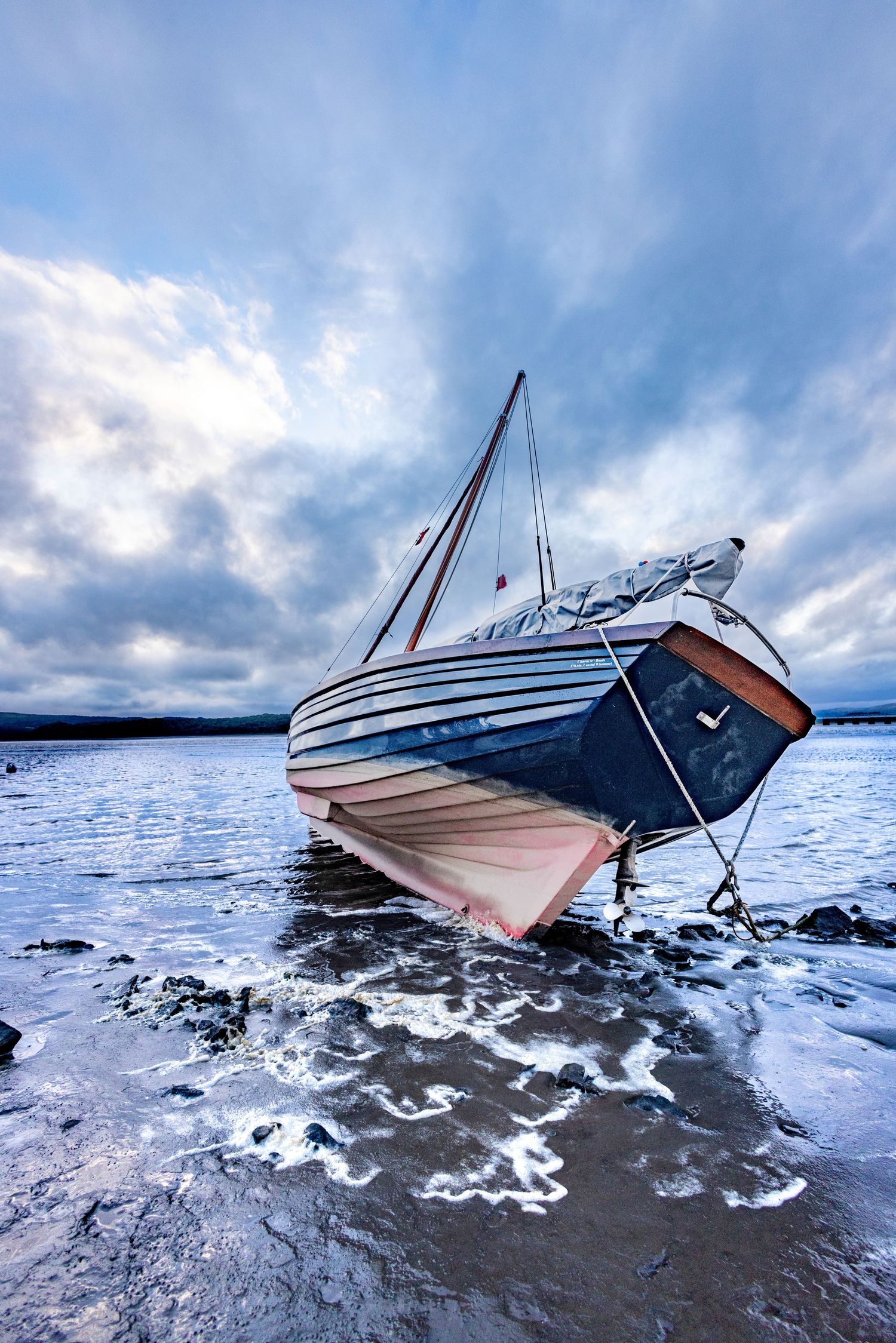 Weathered blue and white sailboat anchored on a rocky tidal flat under stormy clouds.