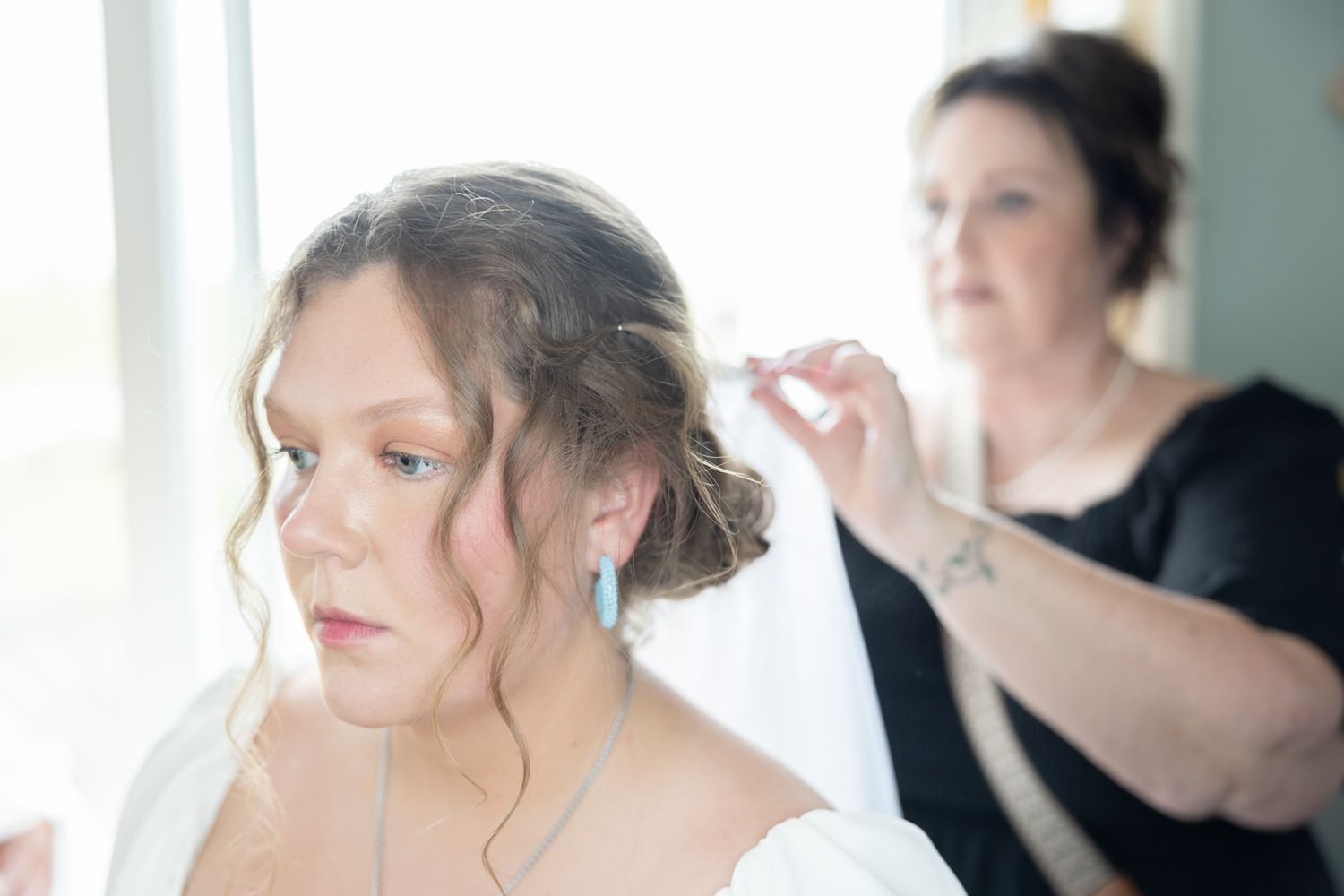 Bride having her hair styled by a woman in soft natural window light before wedding.