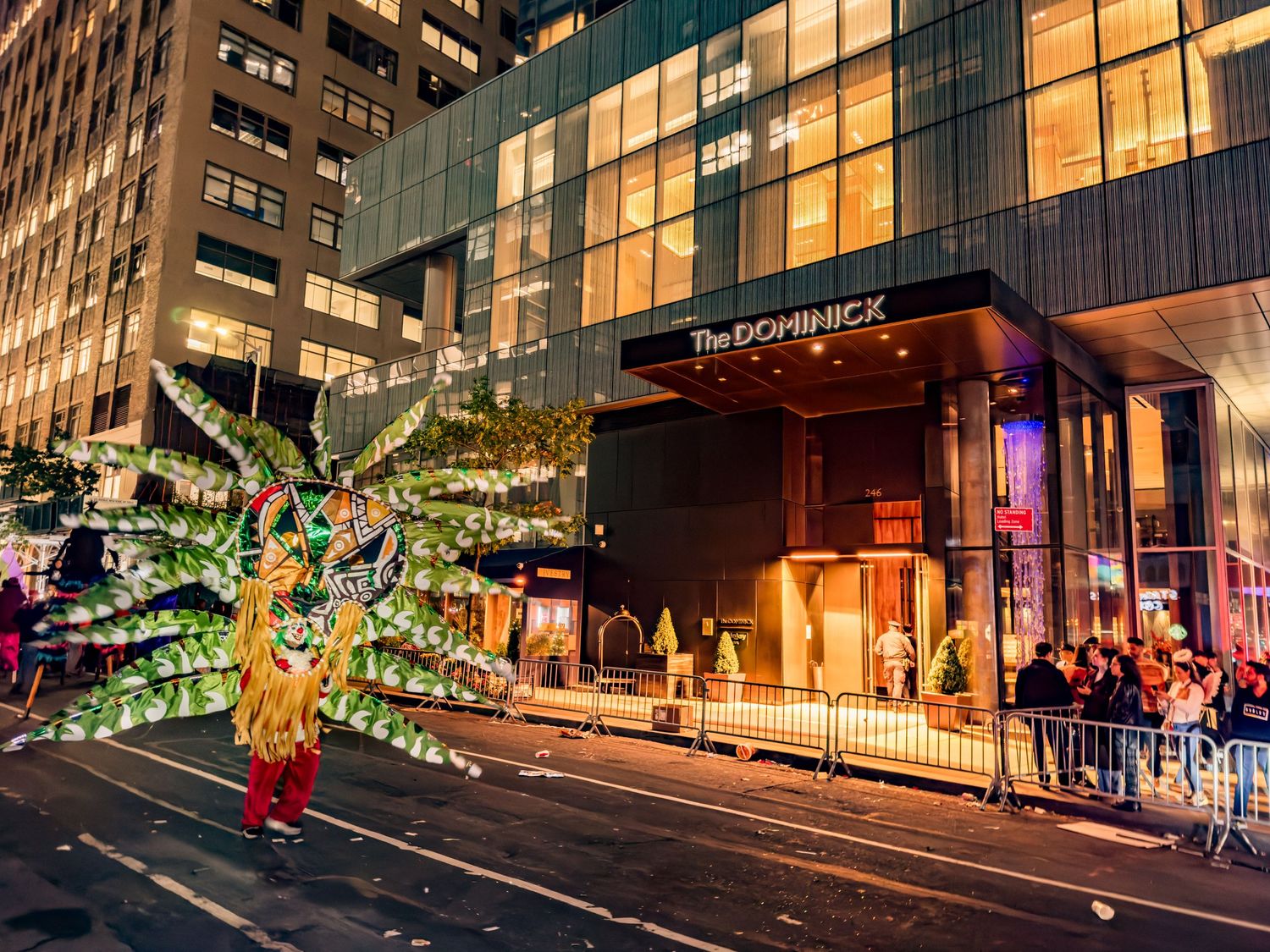 Colorful parade performer in feathered costume outside The Dominick Hotel at night.