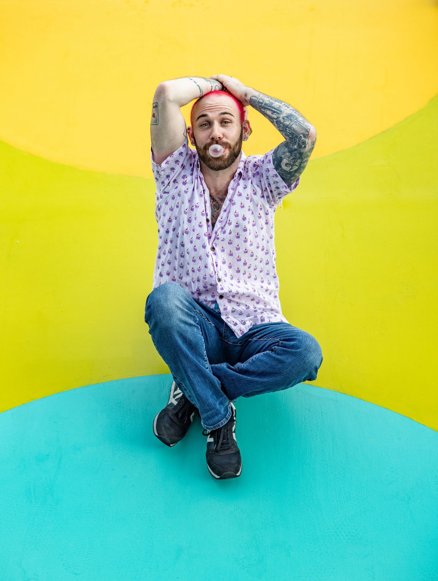 Tattooed man with pink hair crouching on colorful floor against bright yellow and teal background.