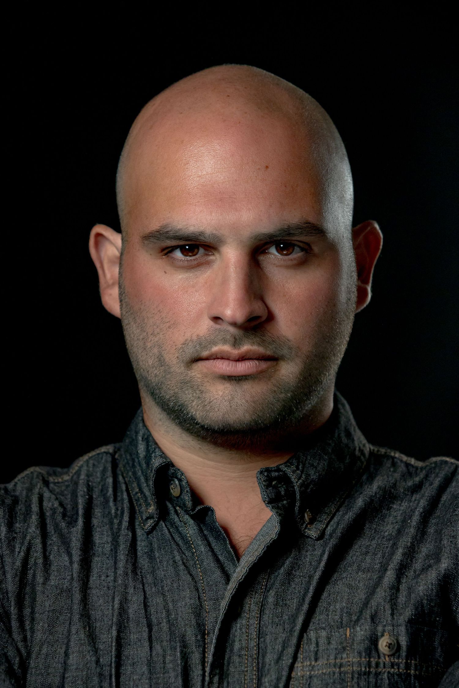 Bald white man with stubble beard wearing dark shirt against black background with serious expression.
