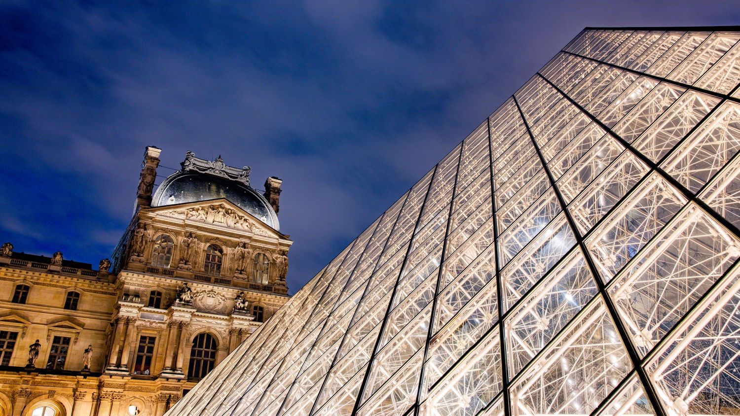 Louvre Museum glass pyramid and historic palace facade illuminated against a dramatic blue twilight sky in Paris.