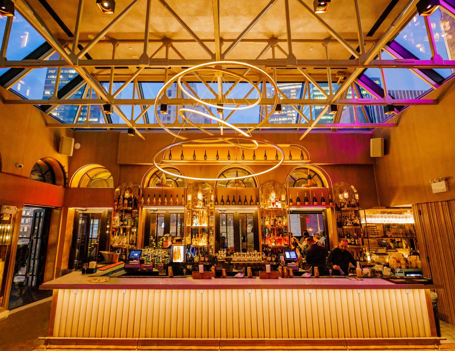 Elegant bar interior with golden lighting, circular chandelier, and colorful bottles beneath a glass ceiling.
