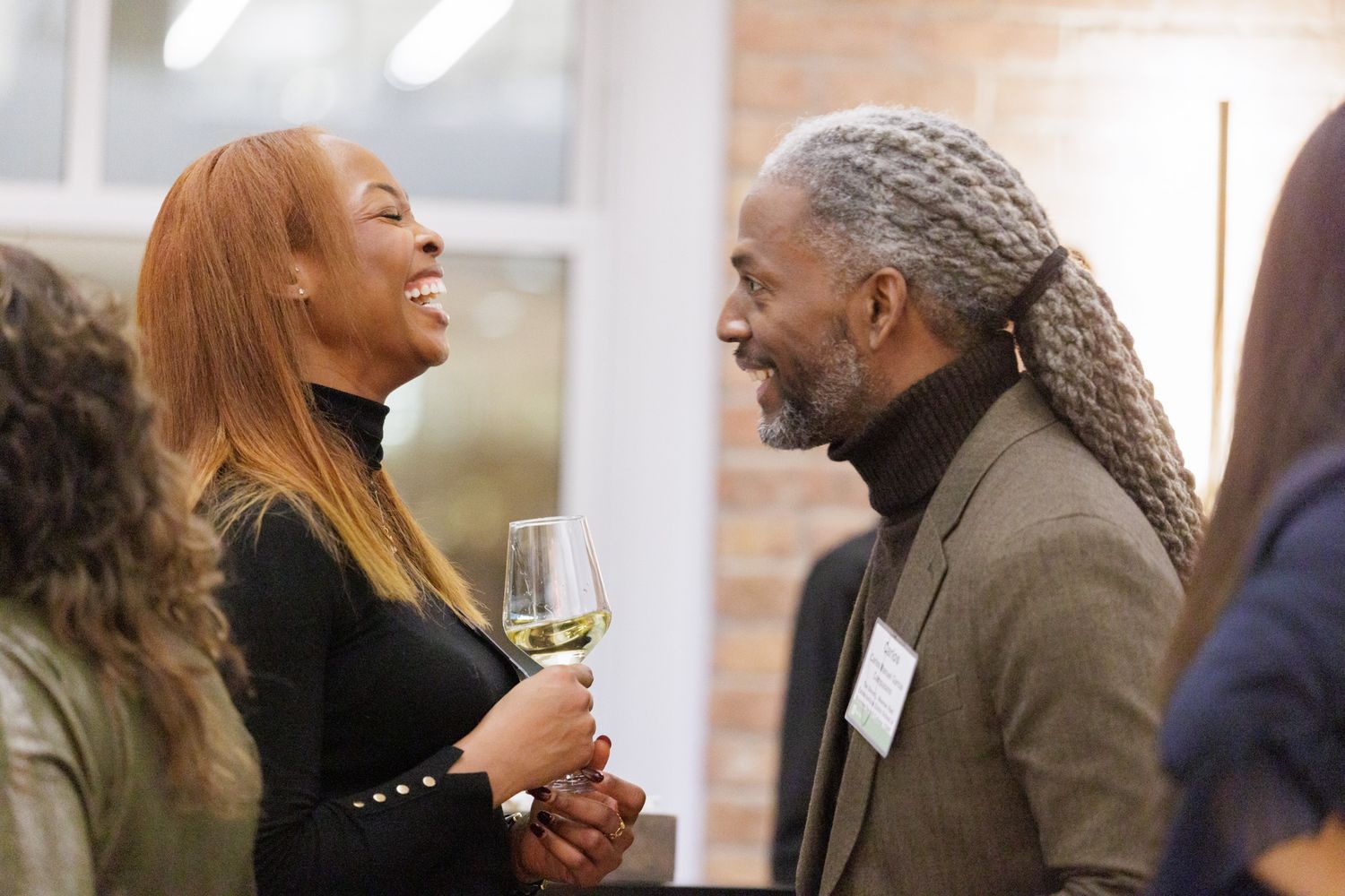 Man and woman laughing together at a social networking event, woman holding a glass of white wine.