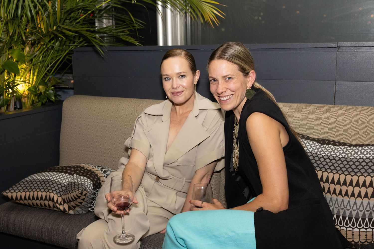 Two women sitting together on a sofa holding wine glasses at a stylish venue.
