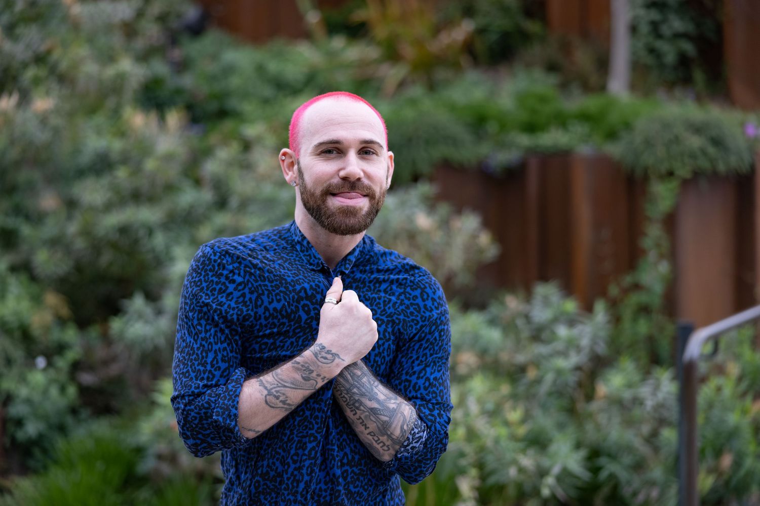 Tattooed man with pink hair in blue patterned shirt crossing arms in lush garden setting.