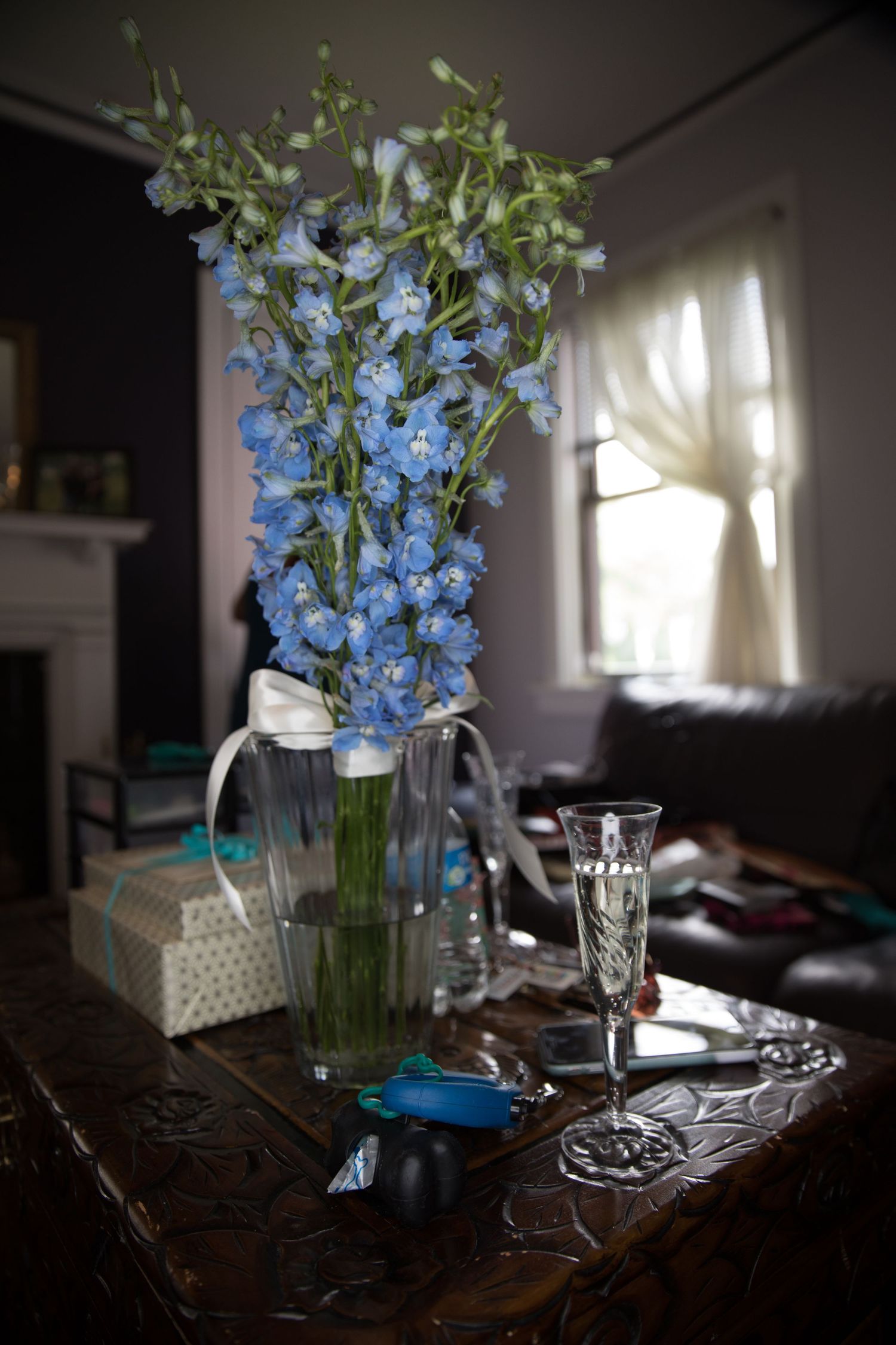 Blue delphinium flowers in glass pitcher vase on ornate wooden table with champagne flute beside it.