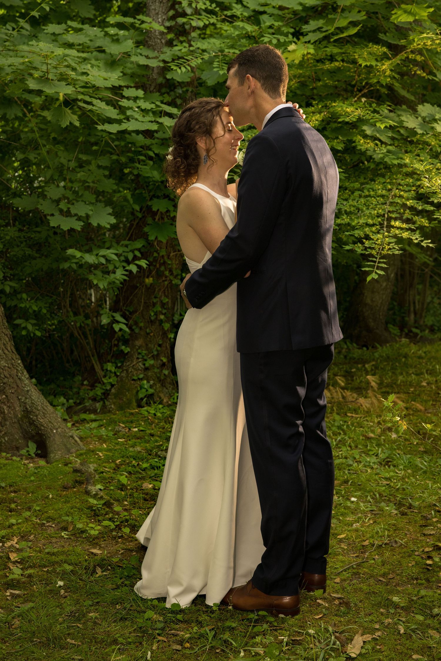 Bride and groom share a romantic embrace in a lush green forest during their outdoor wedding ceremony.