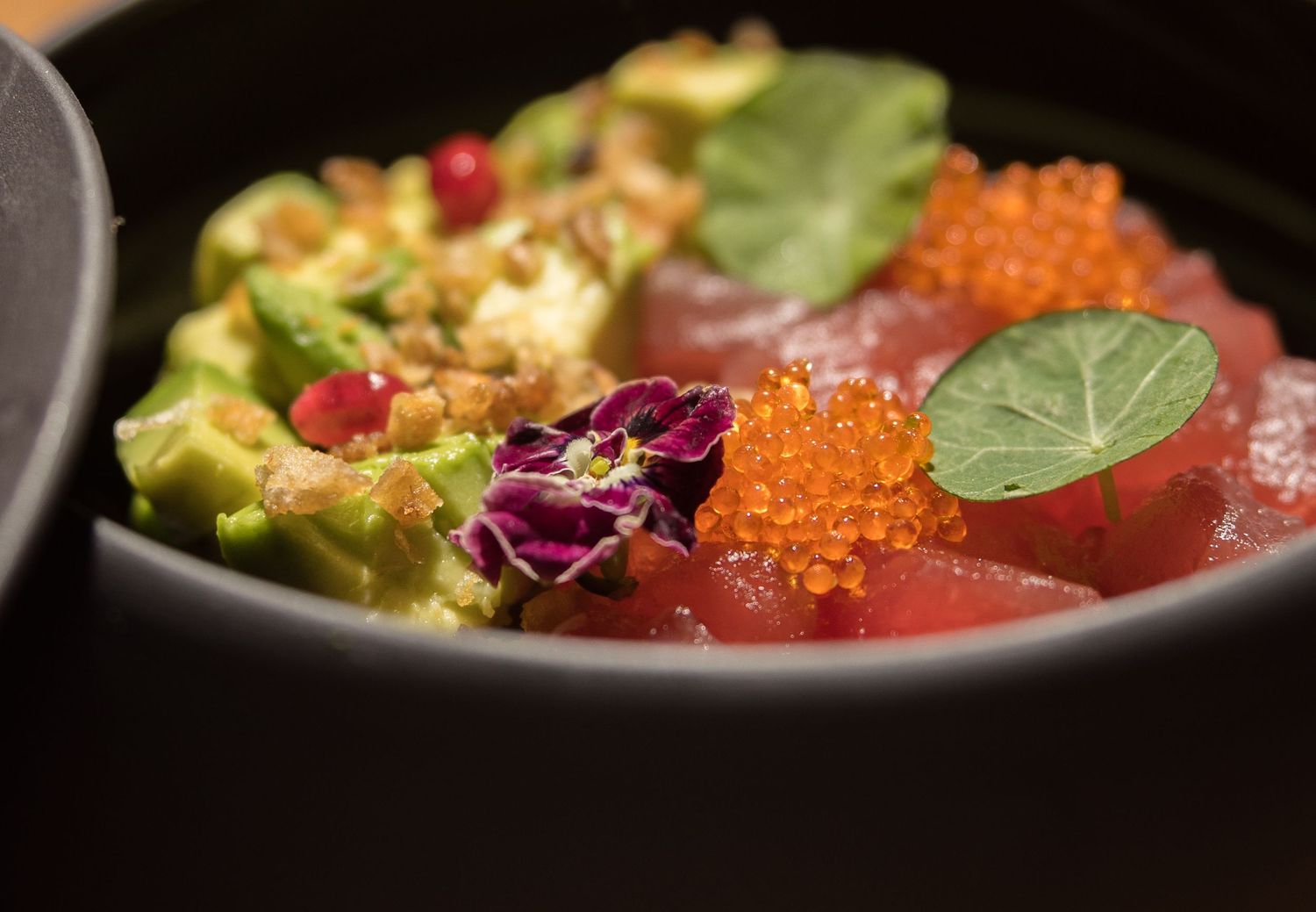 Fresh tuna poke bowl with avocado, tobiko, edible flowers, and microgreens in a dark ceramic bowl.