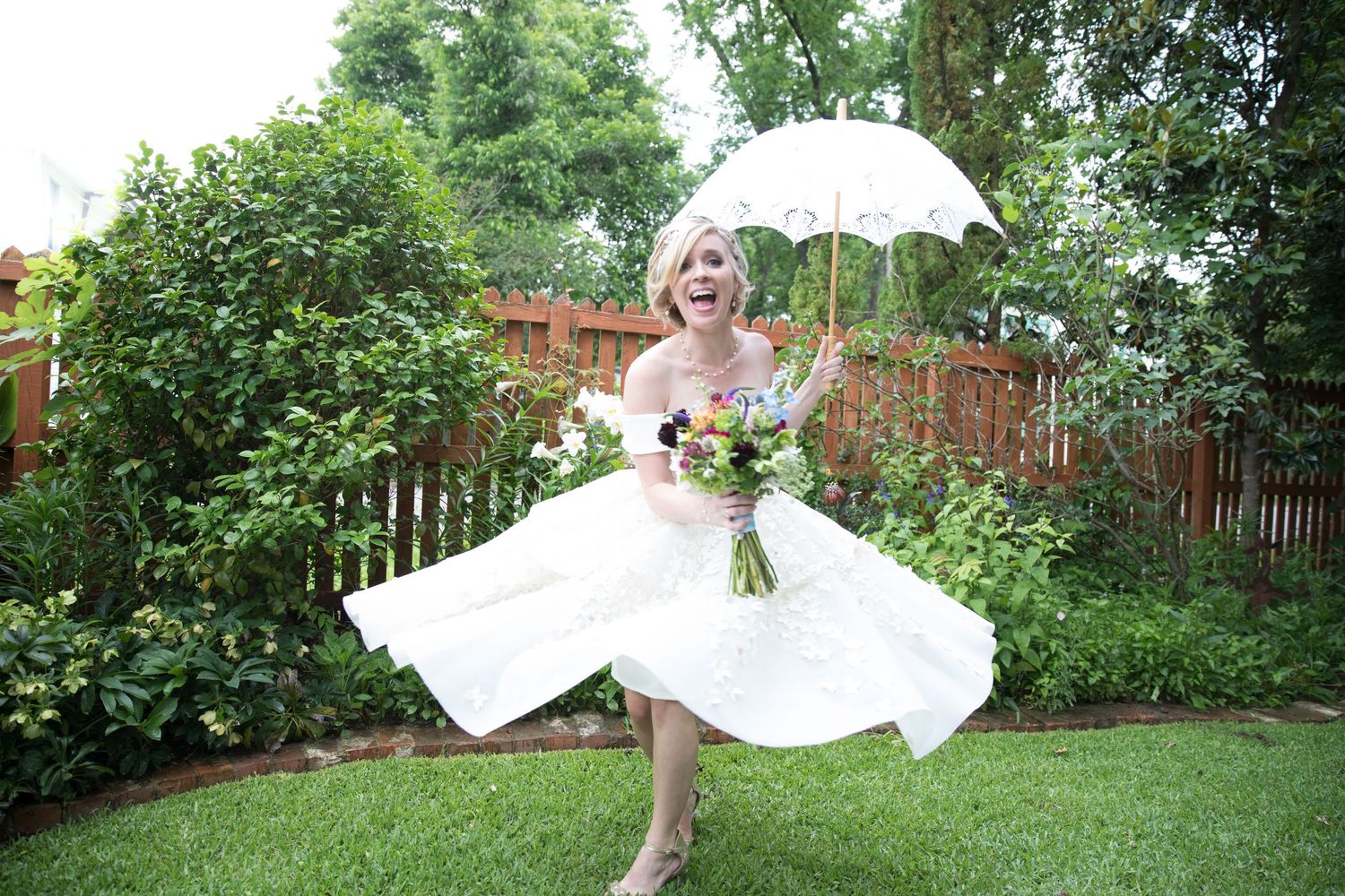 Joyful bride in white dress twirls with umbrella and colorful bouquet in lush green garden.
