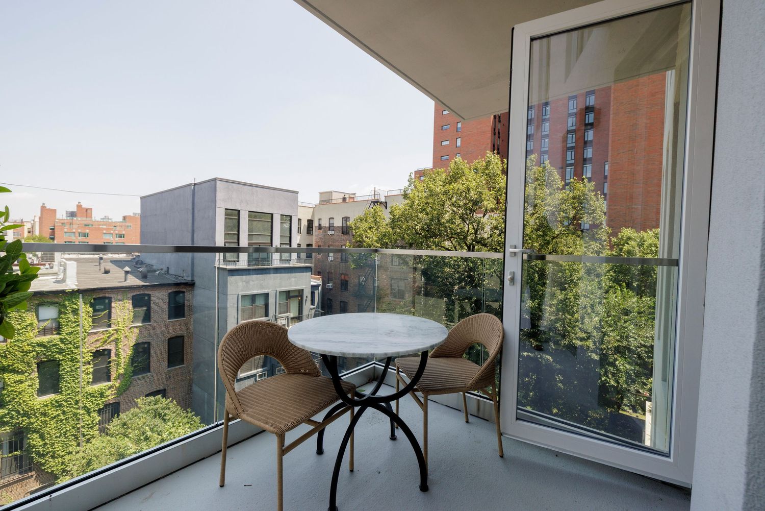 Private apartment balcony with wicker chairs and round table overlooking a lush urban neighborhood.