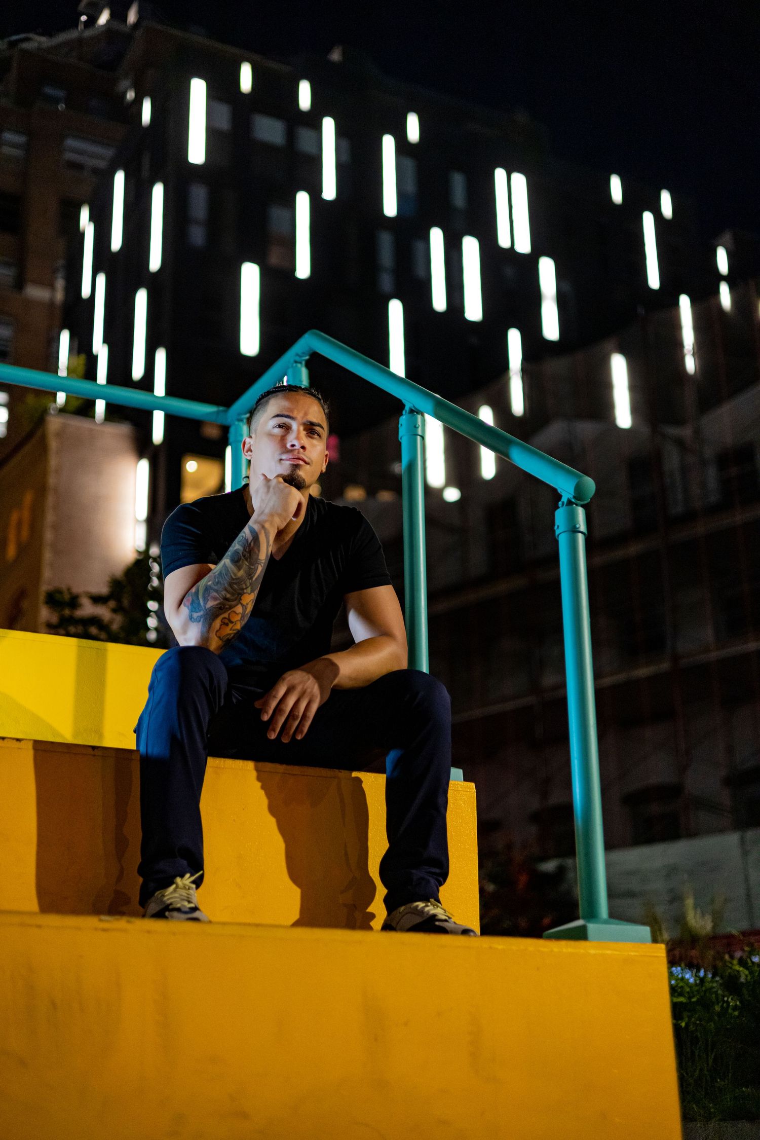 Young man with tattoos sitting on yellow steps at night with illuminated building backdrop.
