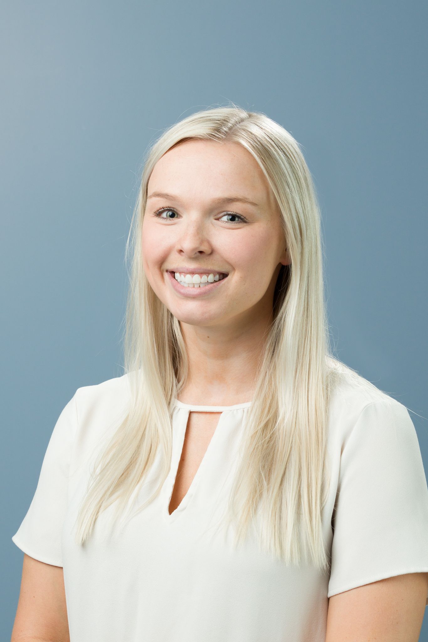 Smiling young blonde woman in a white blouse posing for a professional headshot on blue background.