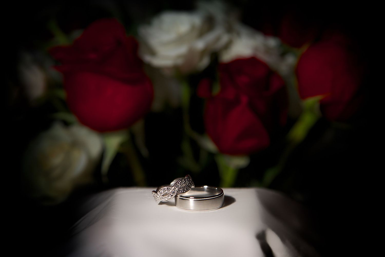Wedding rings displayed on white fabric with red and white roses in the background.