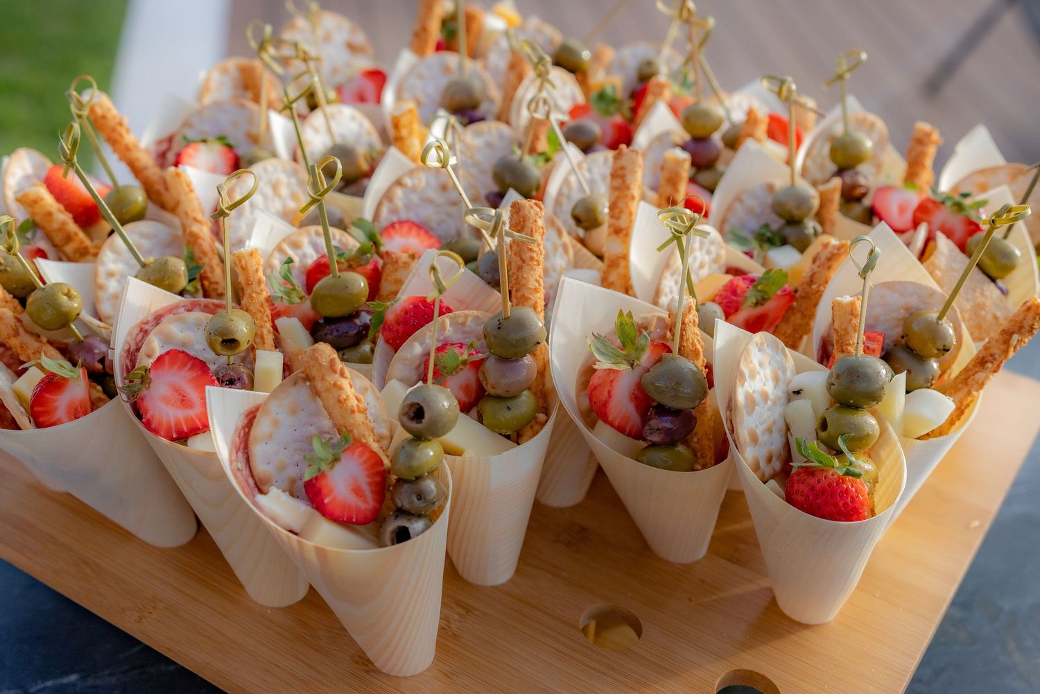 Elegant party appetizer cones filled with fresh strawberries, green olives, and breadsticks arranged on a wooden serving board.
