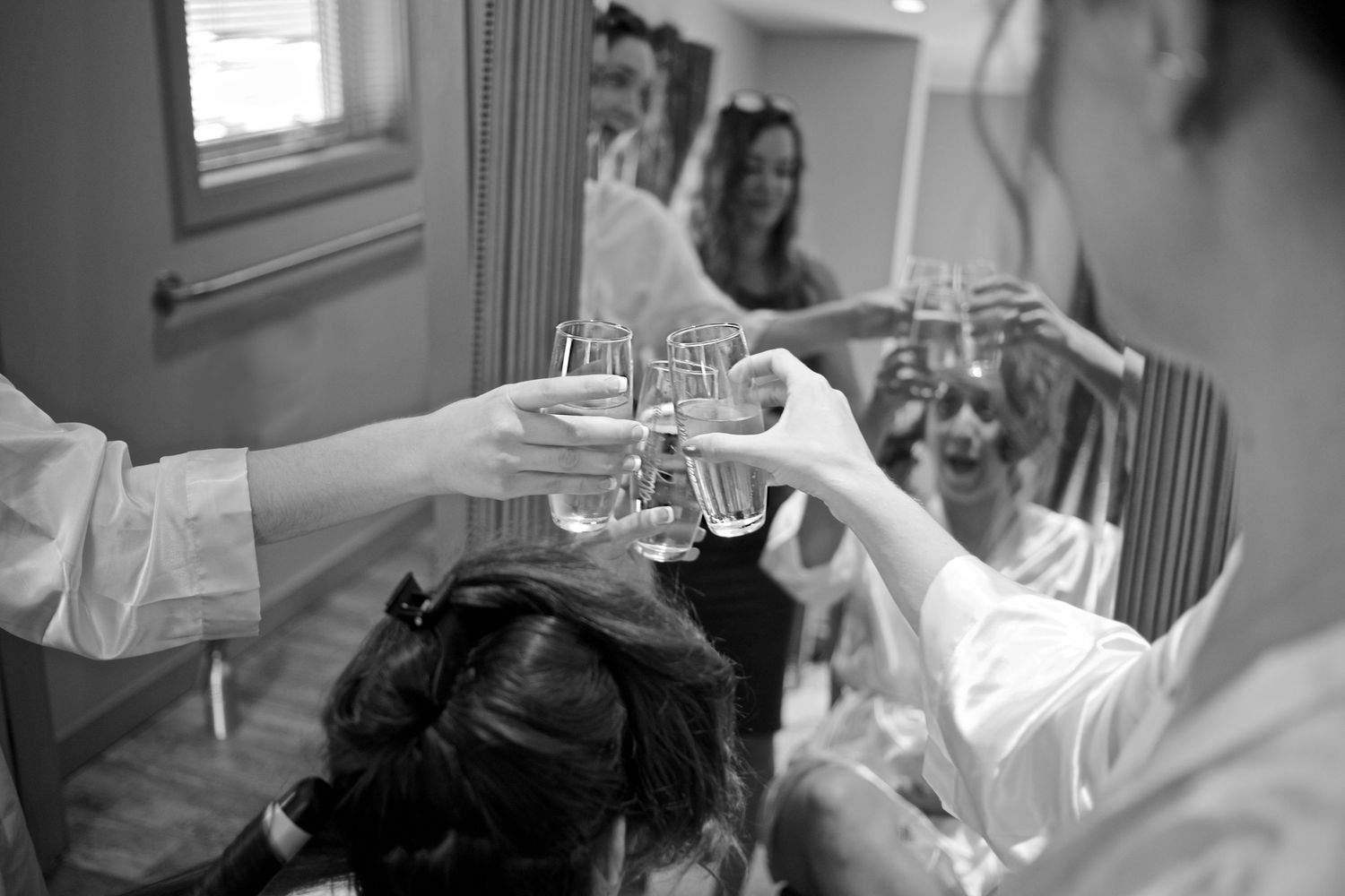 Bridesmaids in white robes toasting with champagne glasses during wedding morning preparations, black and white photo.