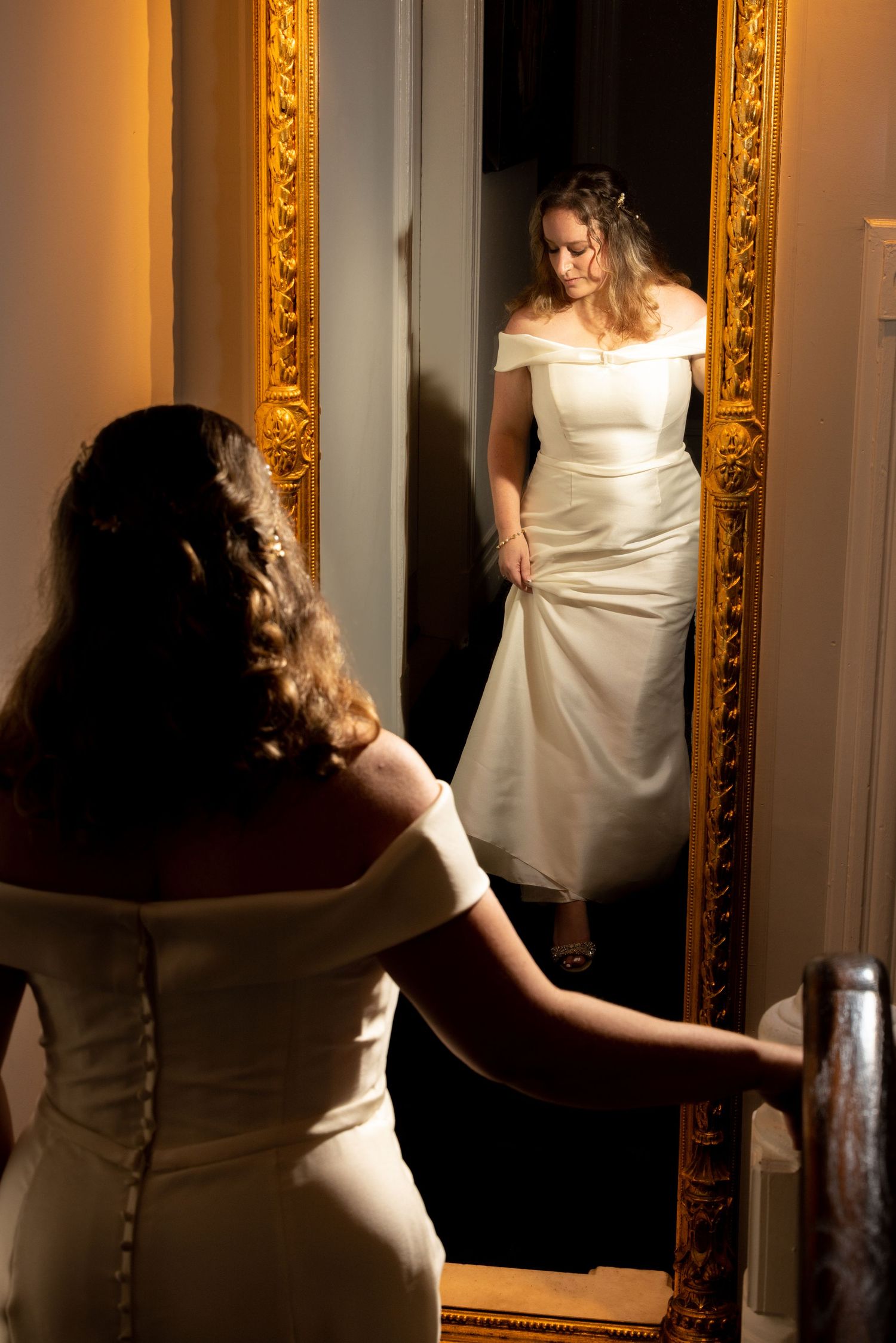 Bride in white off-shoulder gown admiring her reflection in an ornate gold mirror.