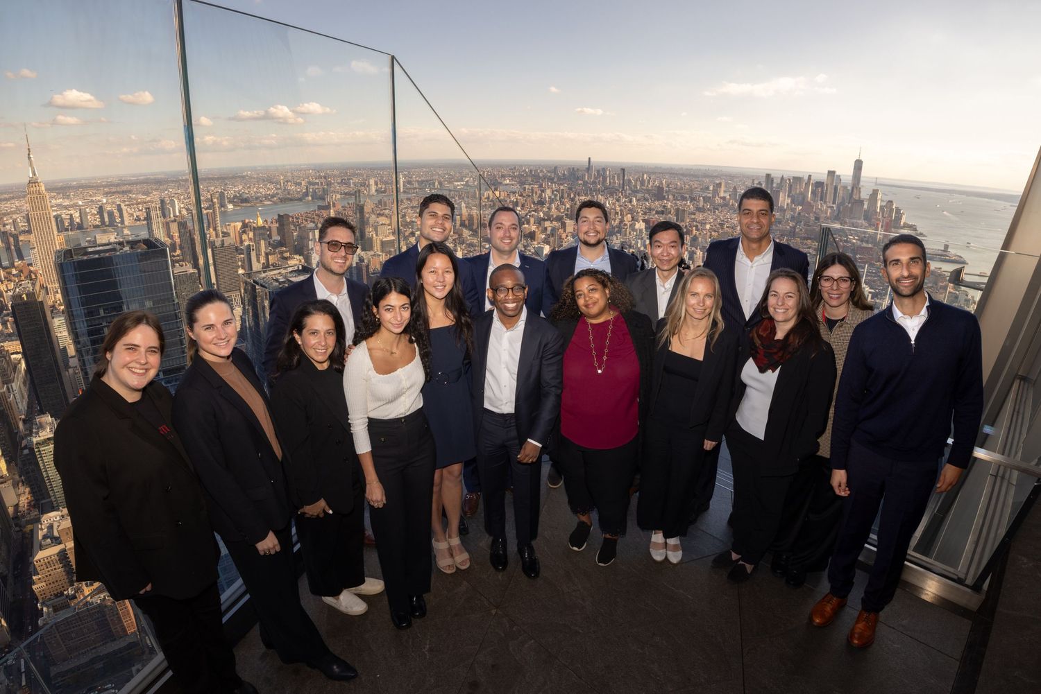 Diverse group of young professionals posing together on a rooftop overlooking a city skyline at dusk.