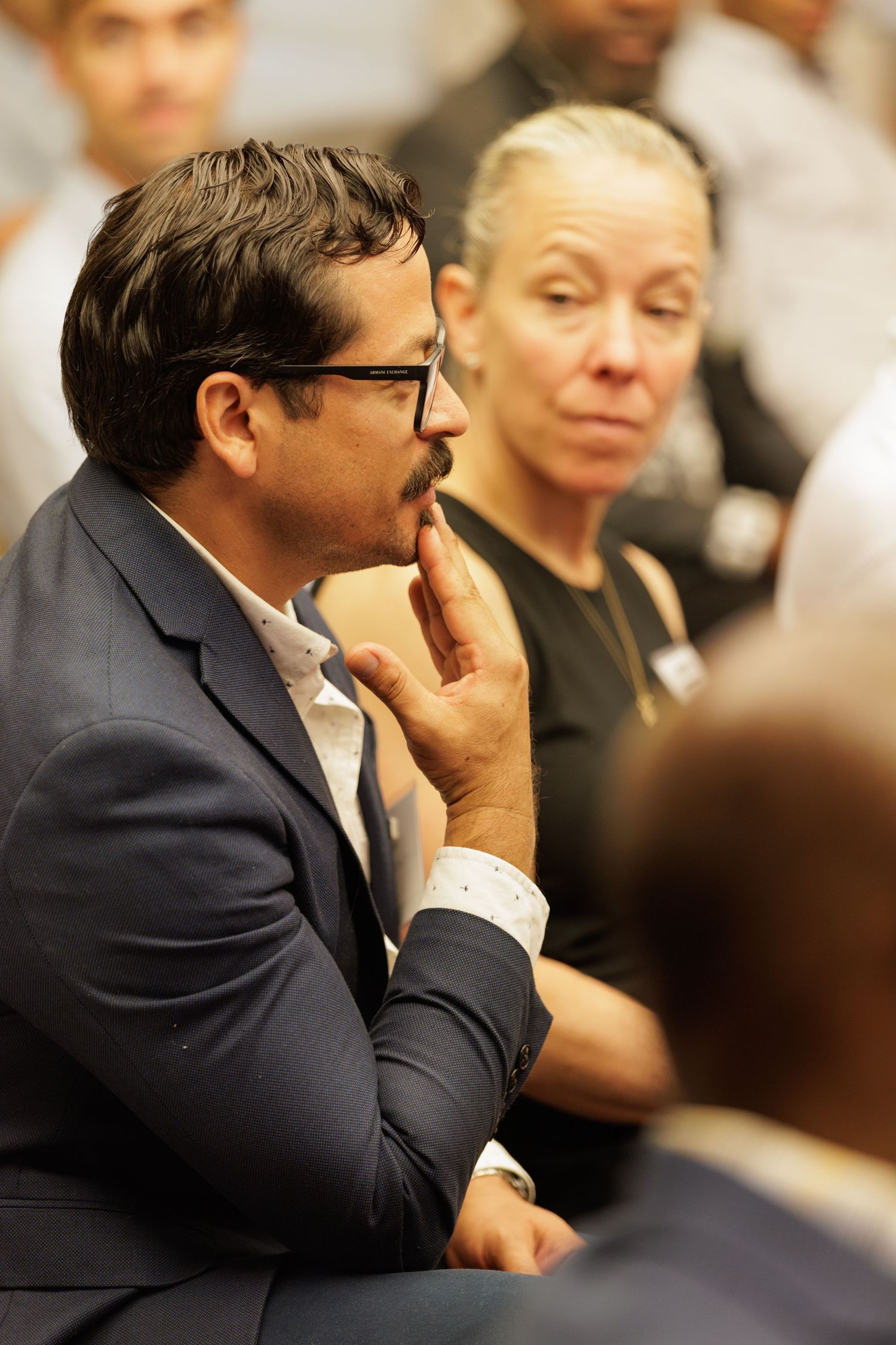 A man in a blue blazer with a mustache listens attentively at a professional conference event.