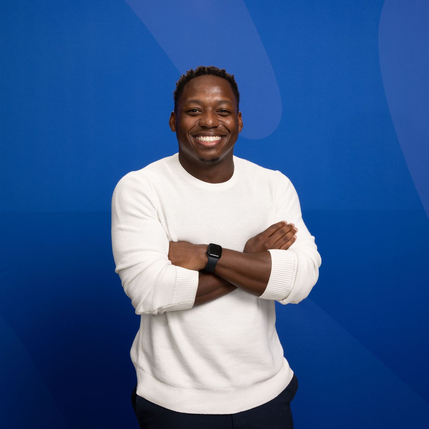Smiling Black man in white sweater with arms crossed against blue background.