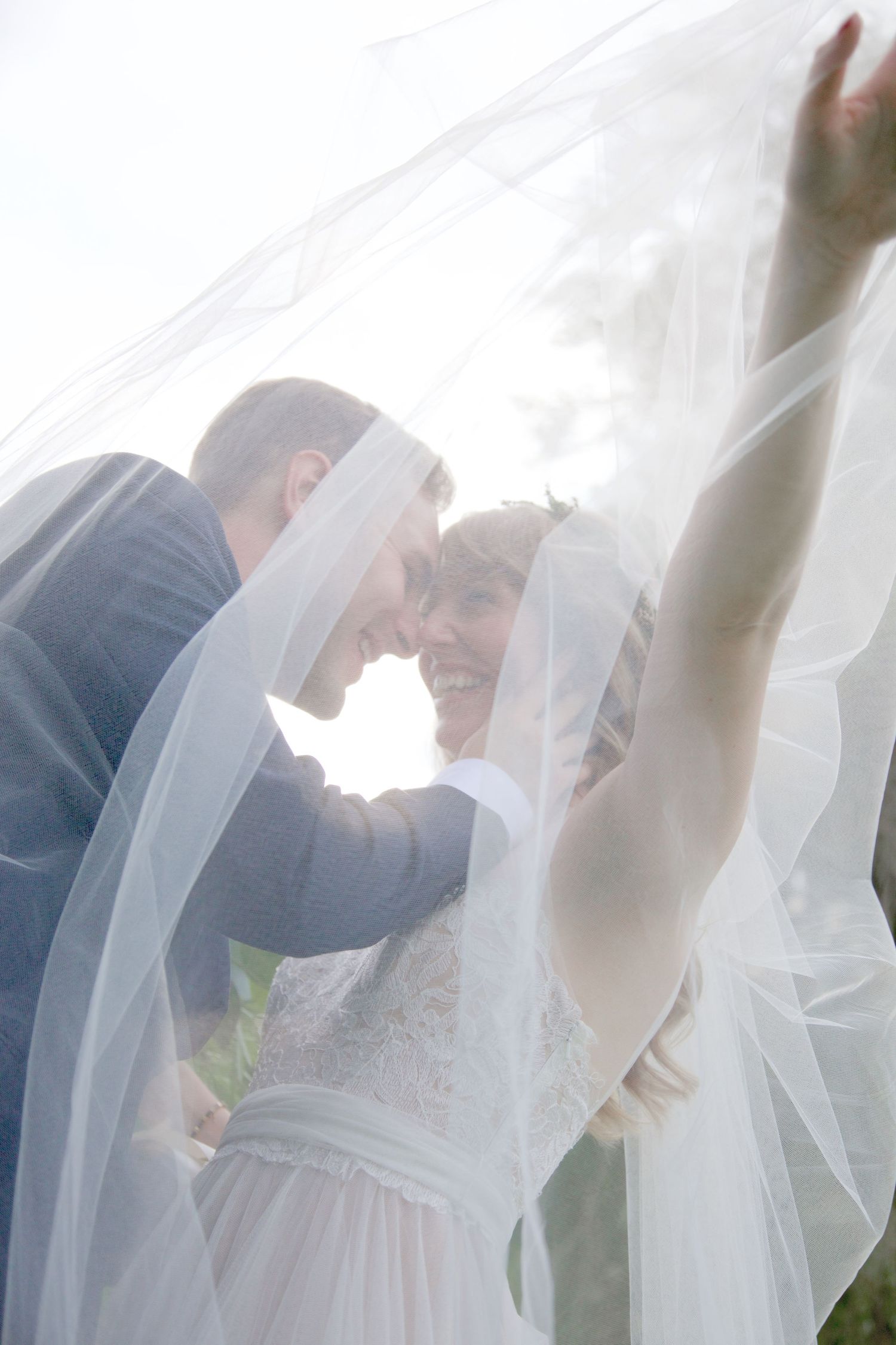 Bride and groom share a romantic moment under flowing white veil with bright backlight.