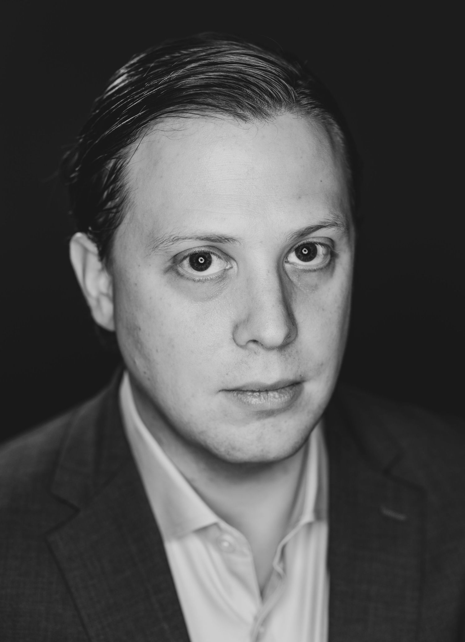 Black and white professional headshot of young white man in suit jacket with slicked back hair.