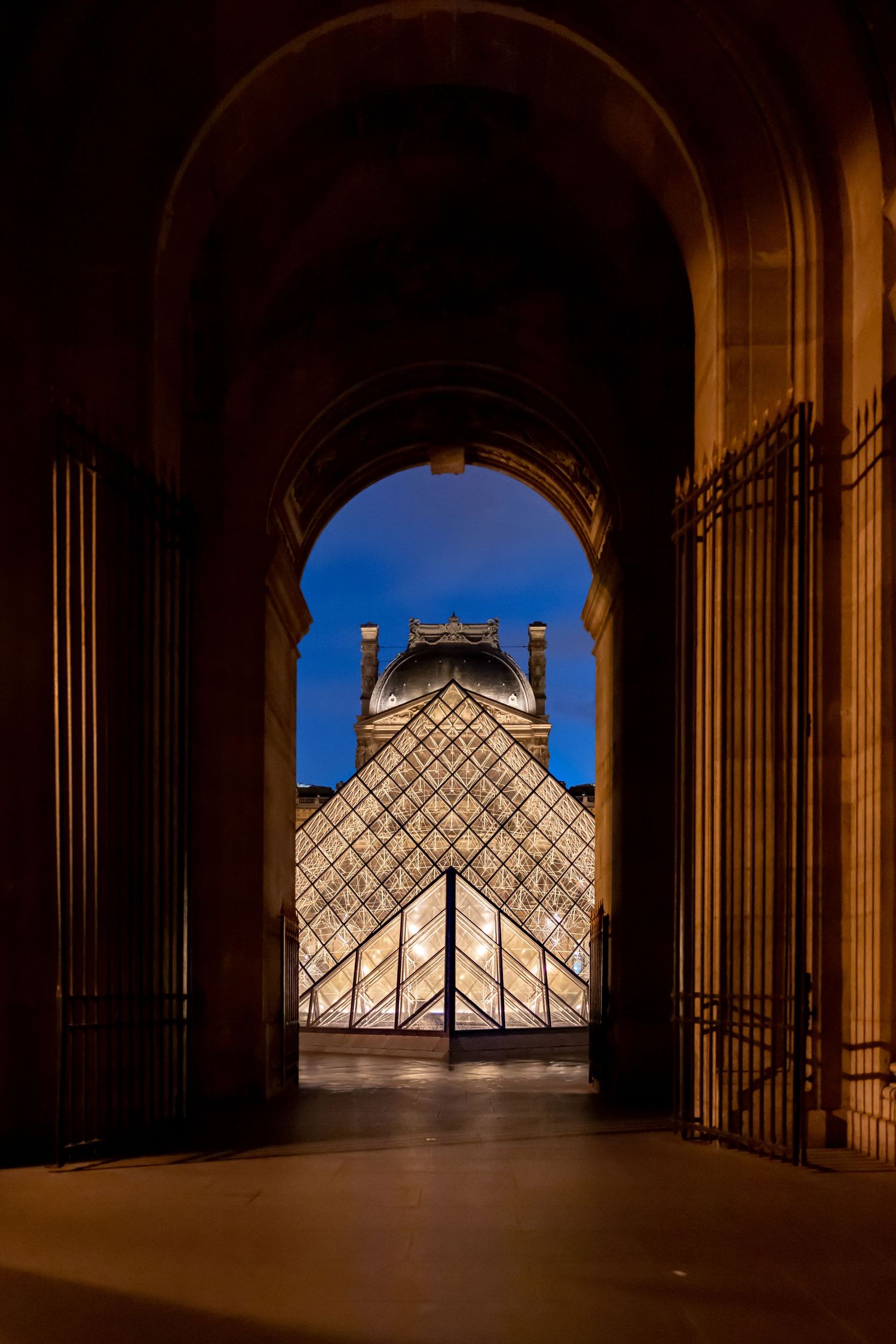 Louvre Museum glass pyramid viewed through a grand stone archway at dusk in Paris, France.