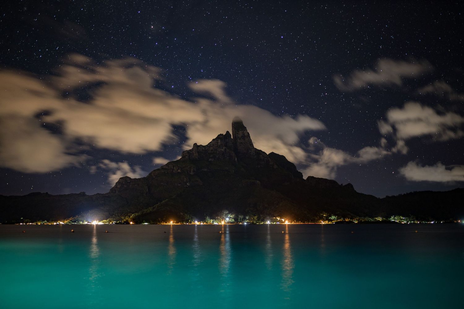 A stunning night sky with stars and clouds illuminates a dramatic mountain island rising above turquoise waters.
