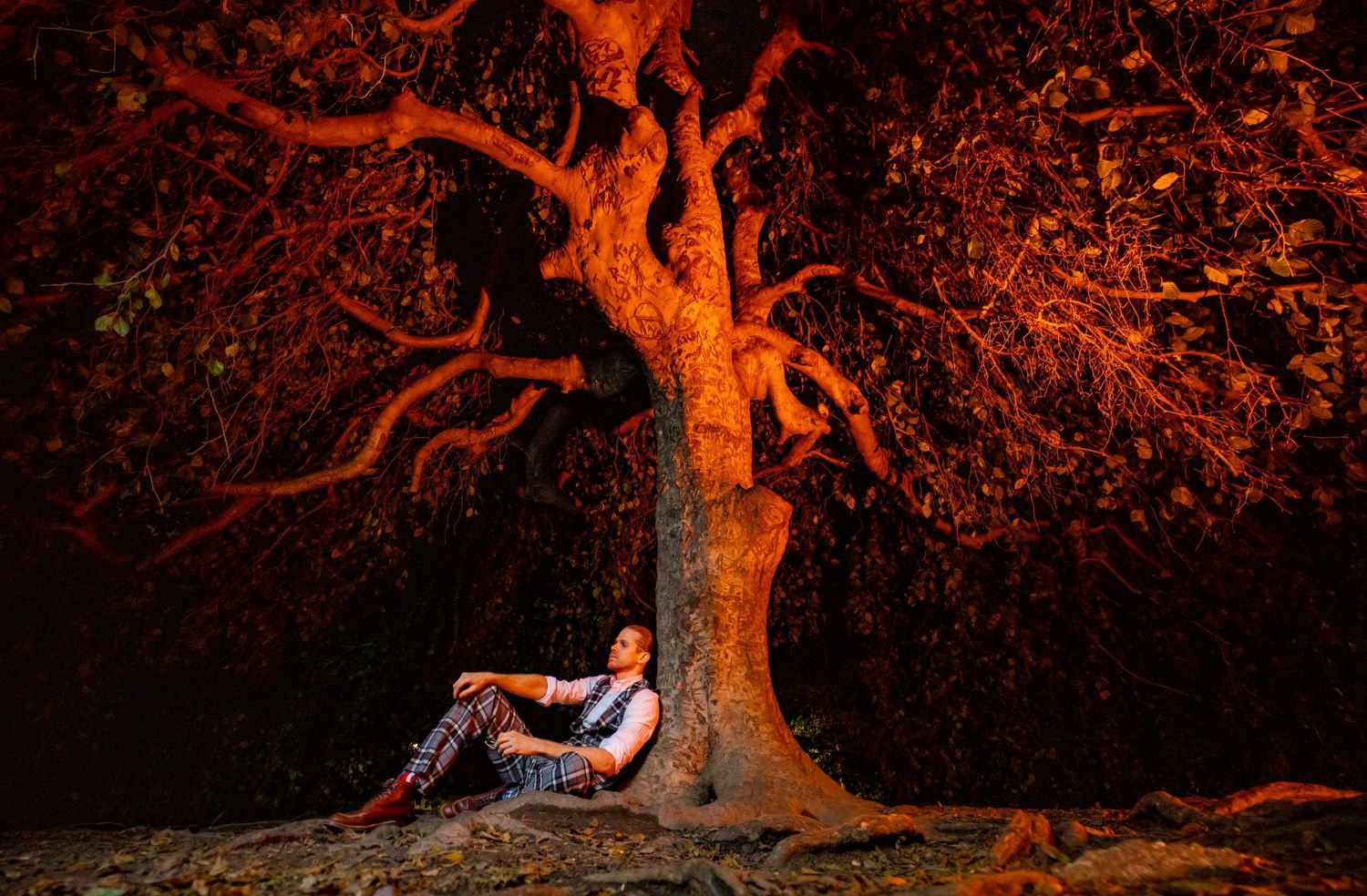 Man in plaid outfit resting against large tree trunk illuminated by dramatic orange light at night.