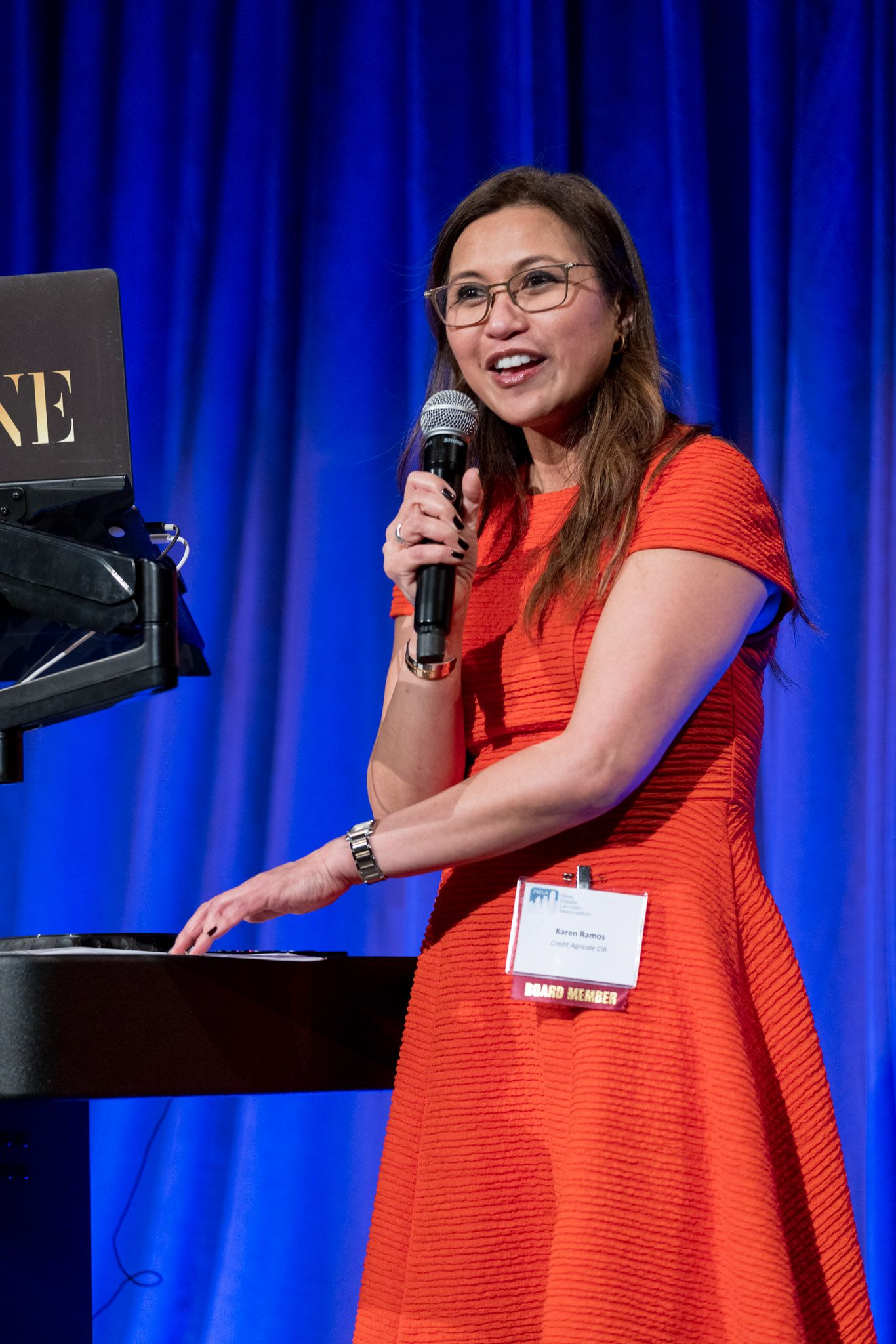Woman in red dress speaking at podium with microphone at a professional conference event.
