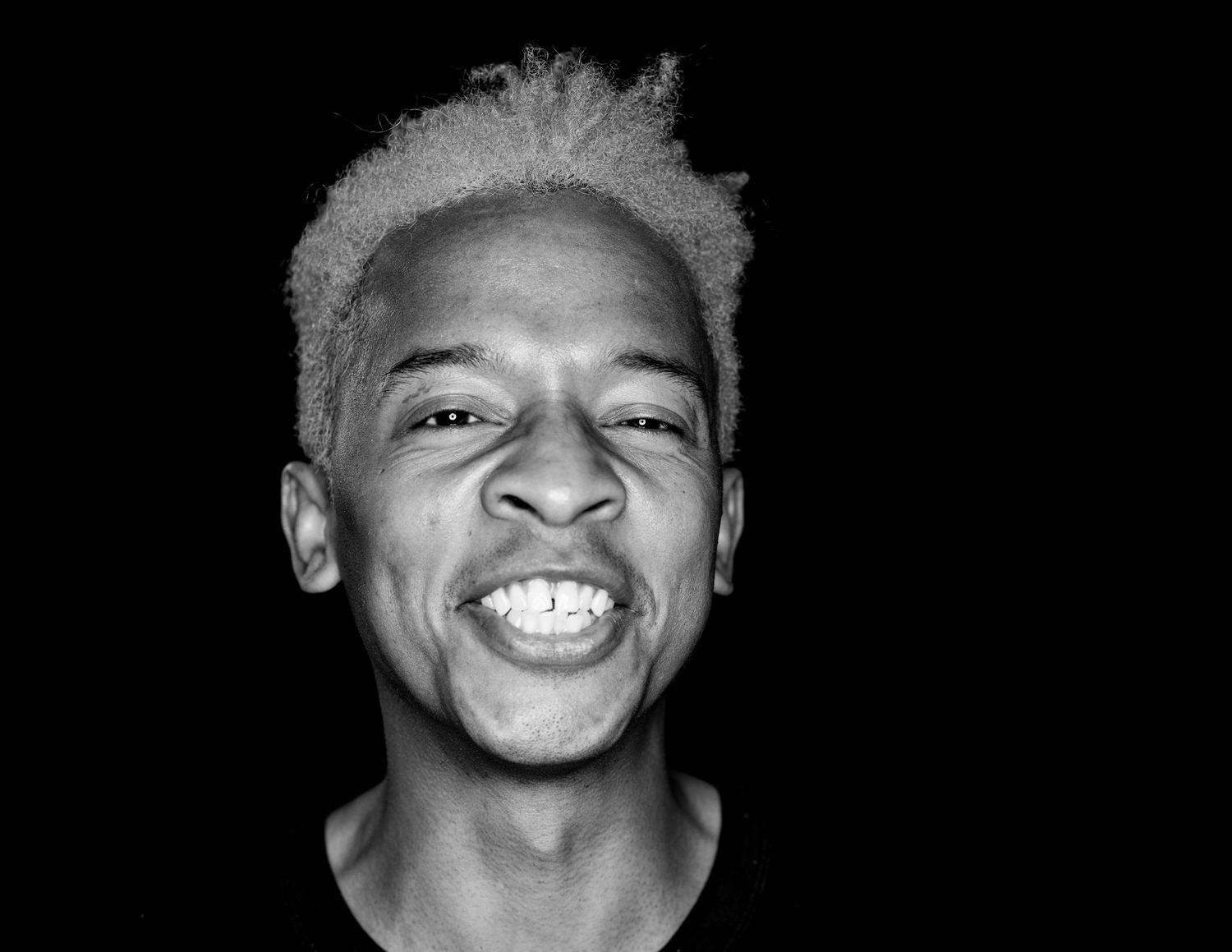 Black and white photo of a young man with blonde hair laughing joyfully against a dark background.