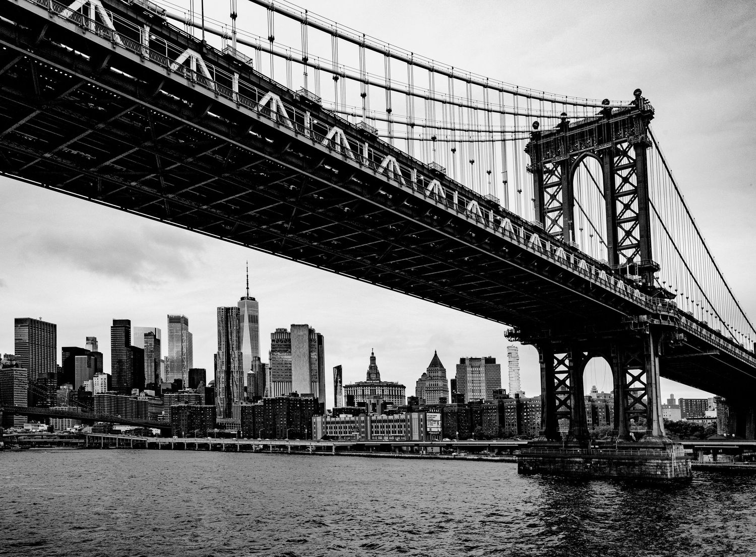 Black and white photo of Manhattan Bridge over East River with NYC skyline and skyscrapers behind.
