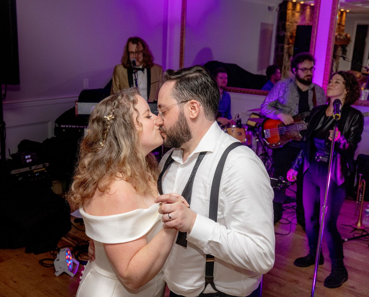 Bride and groom share a romantic first dance kiss while a live band performs in purple-lit reception hall.